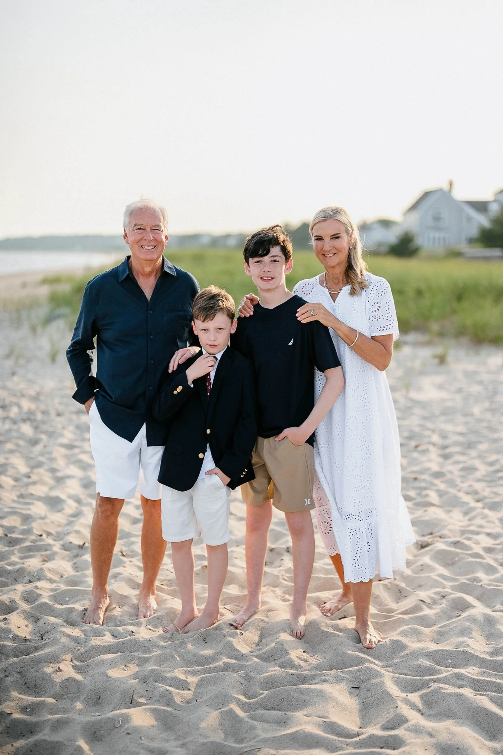 A family of five standing barefoot on the beach with houses in the background, smiling at the camera during sunset.