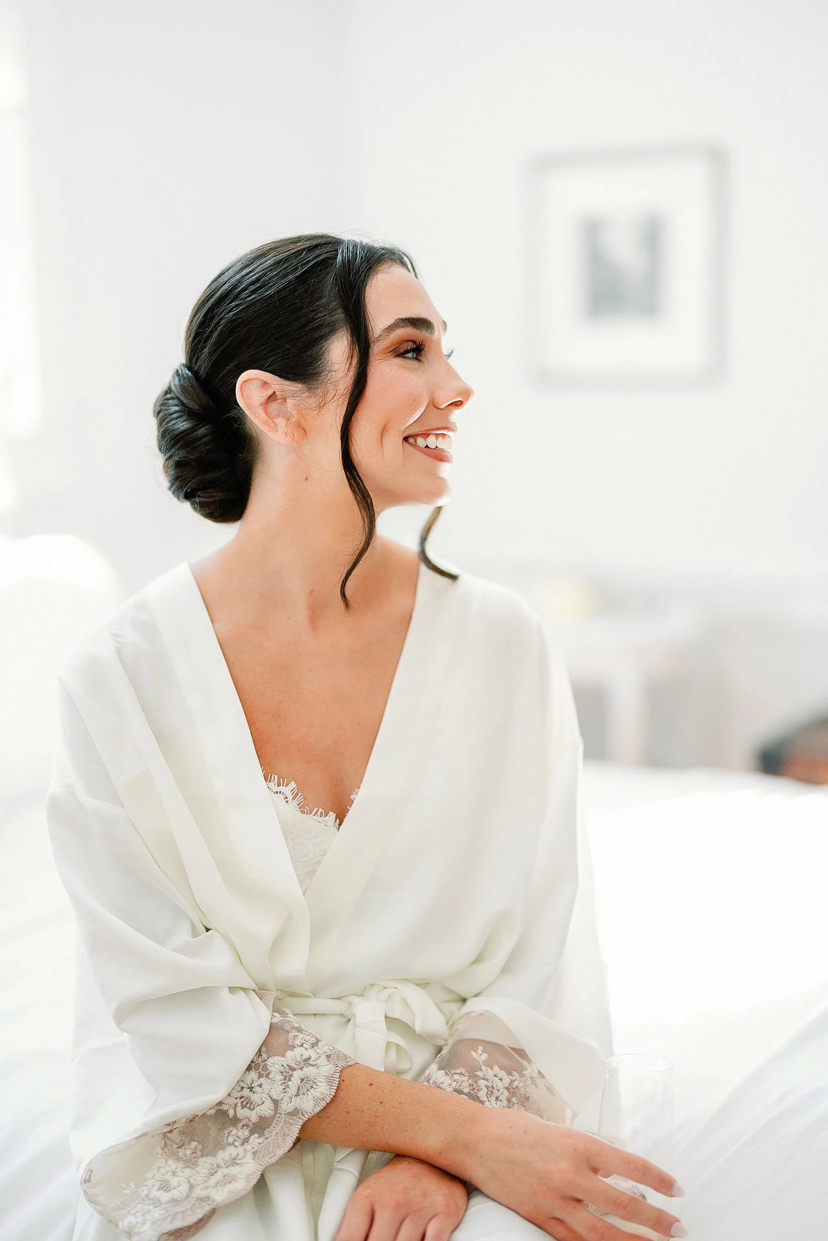 A woman in a white robe with lace trim sits on a bed, smiling and looking to the side in a bright, minimal bedroom.