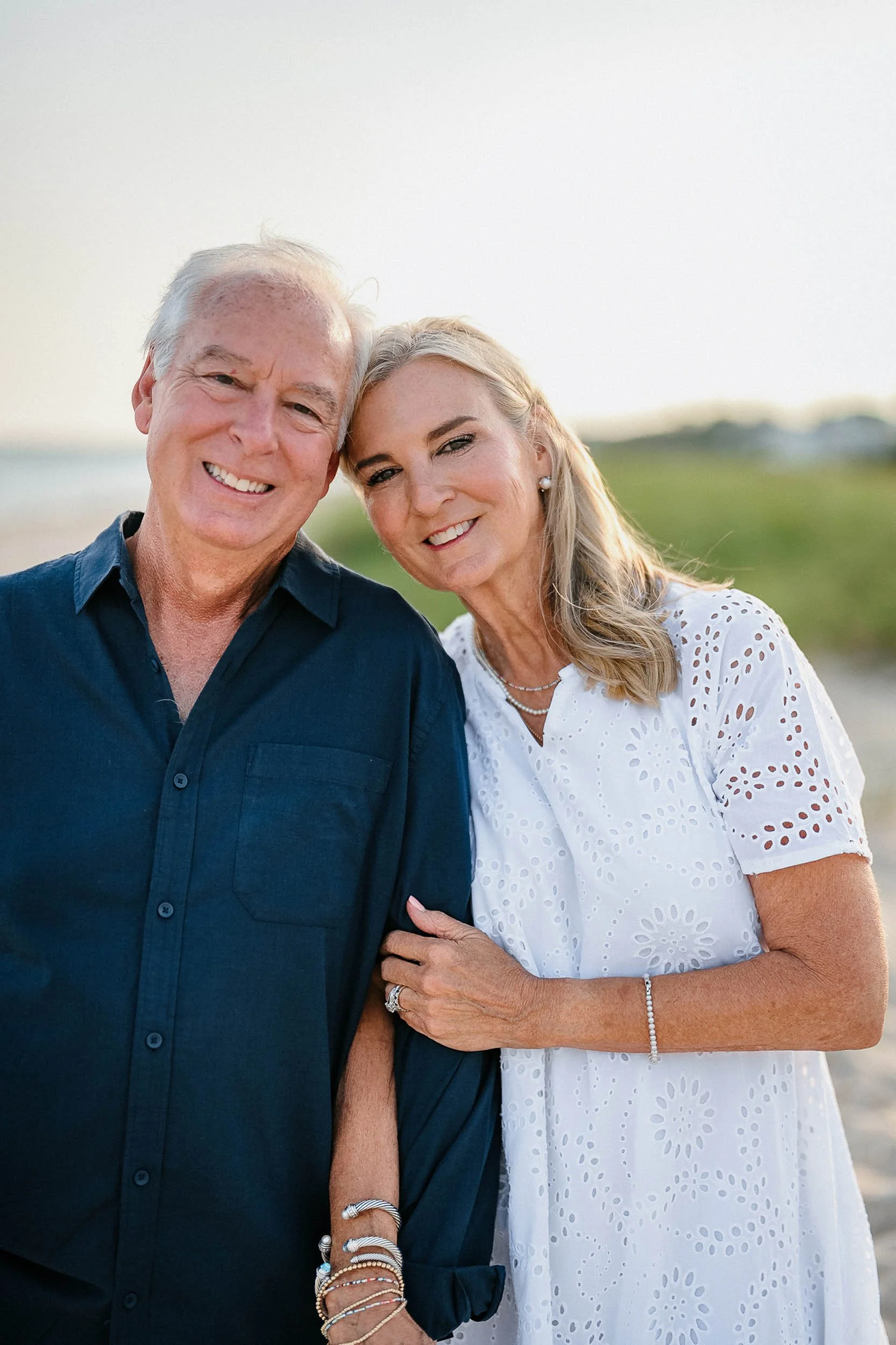 Senior couple smiling and leaning close together outdoors at a beach or park during sunset, dressed casually.