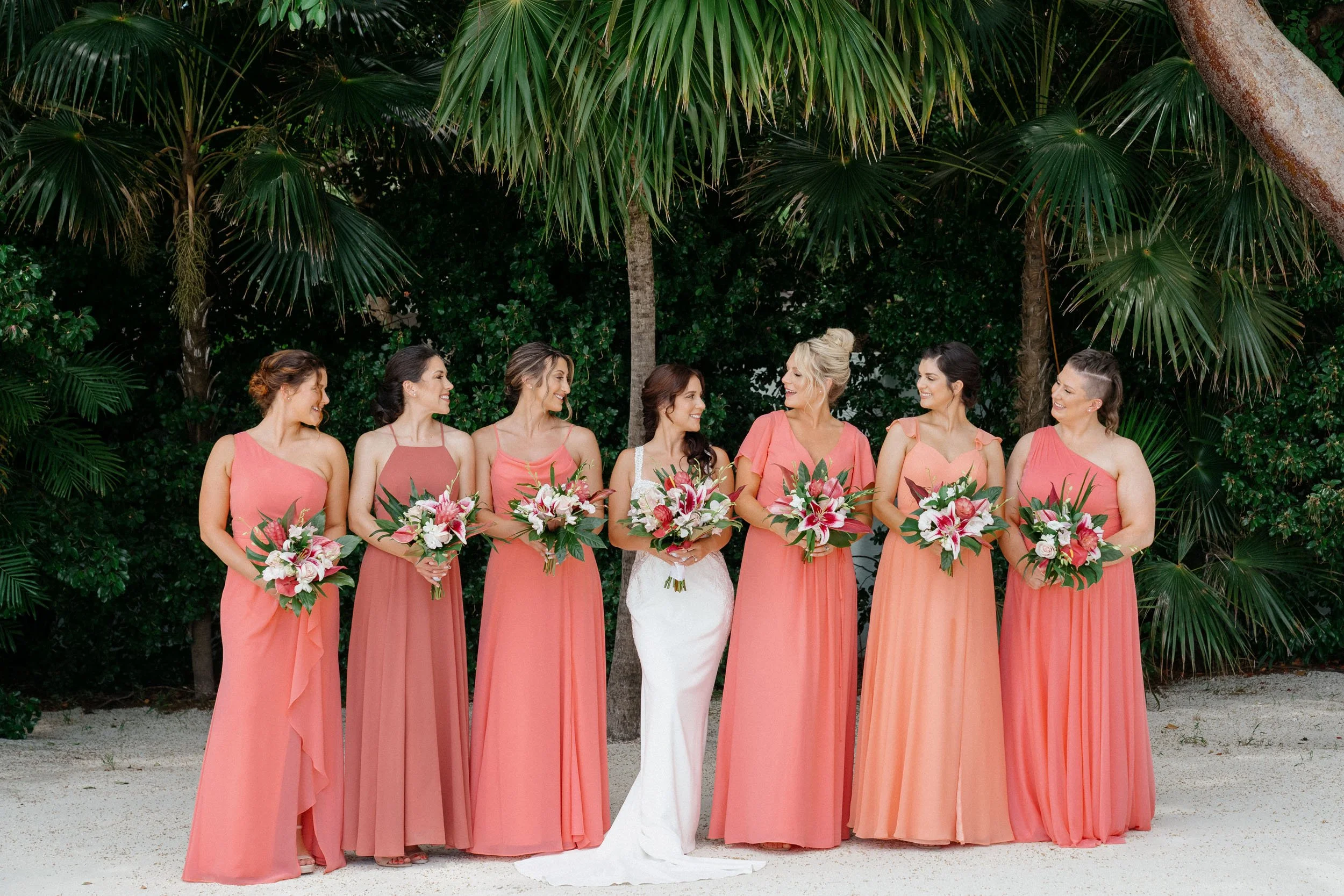 Bride in white dress surrounded by six bridesmaids in pink dresses, all holding bouquets, standing outdoors under palm trees.