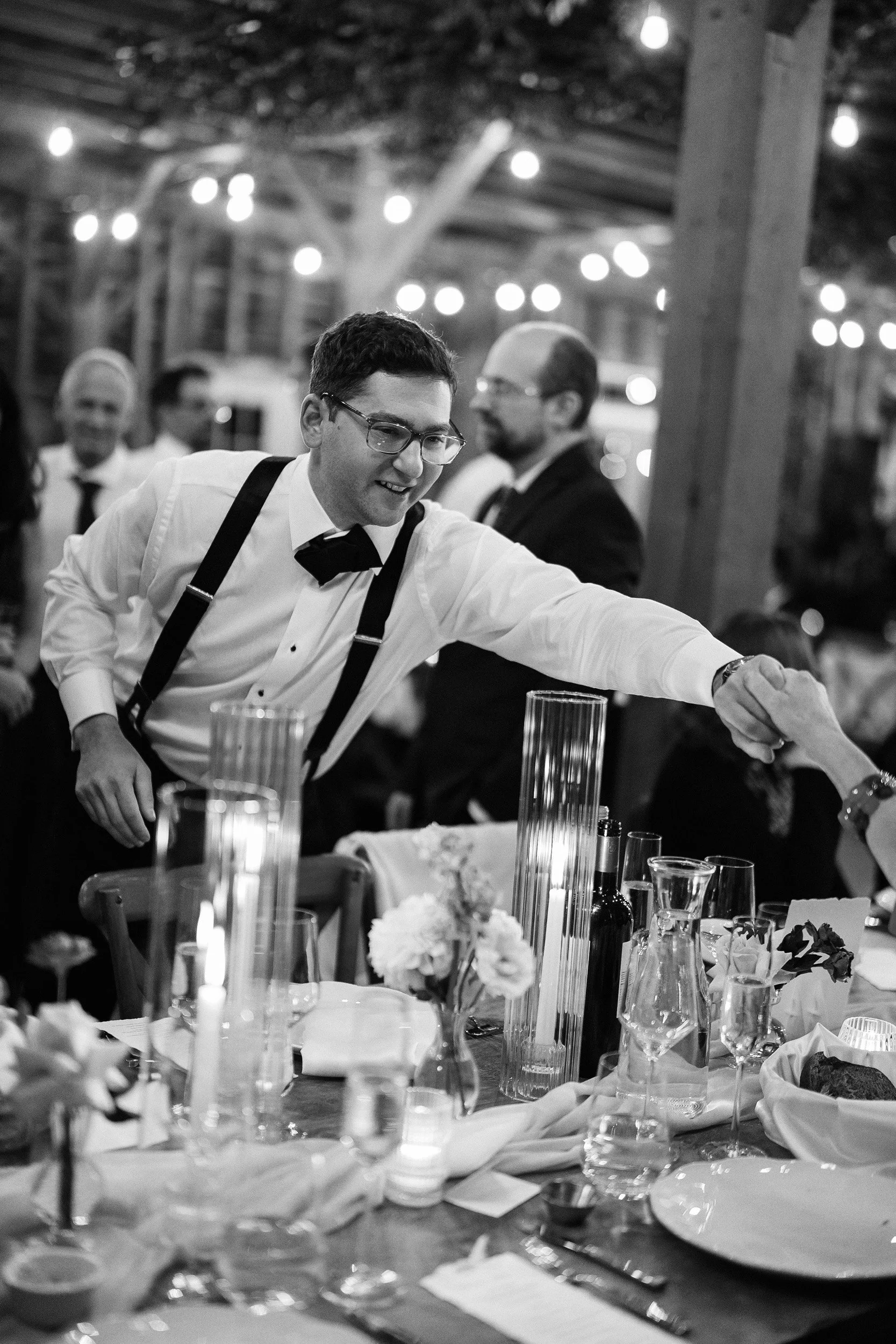 A black and white photo of a man in formal attire, including a bowtie and suspenders, reaching across a decorated dinner table at a social event or celebration.