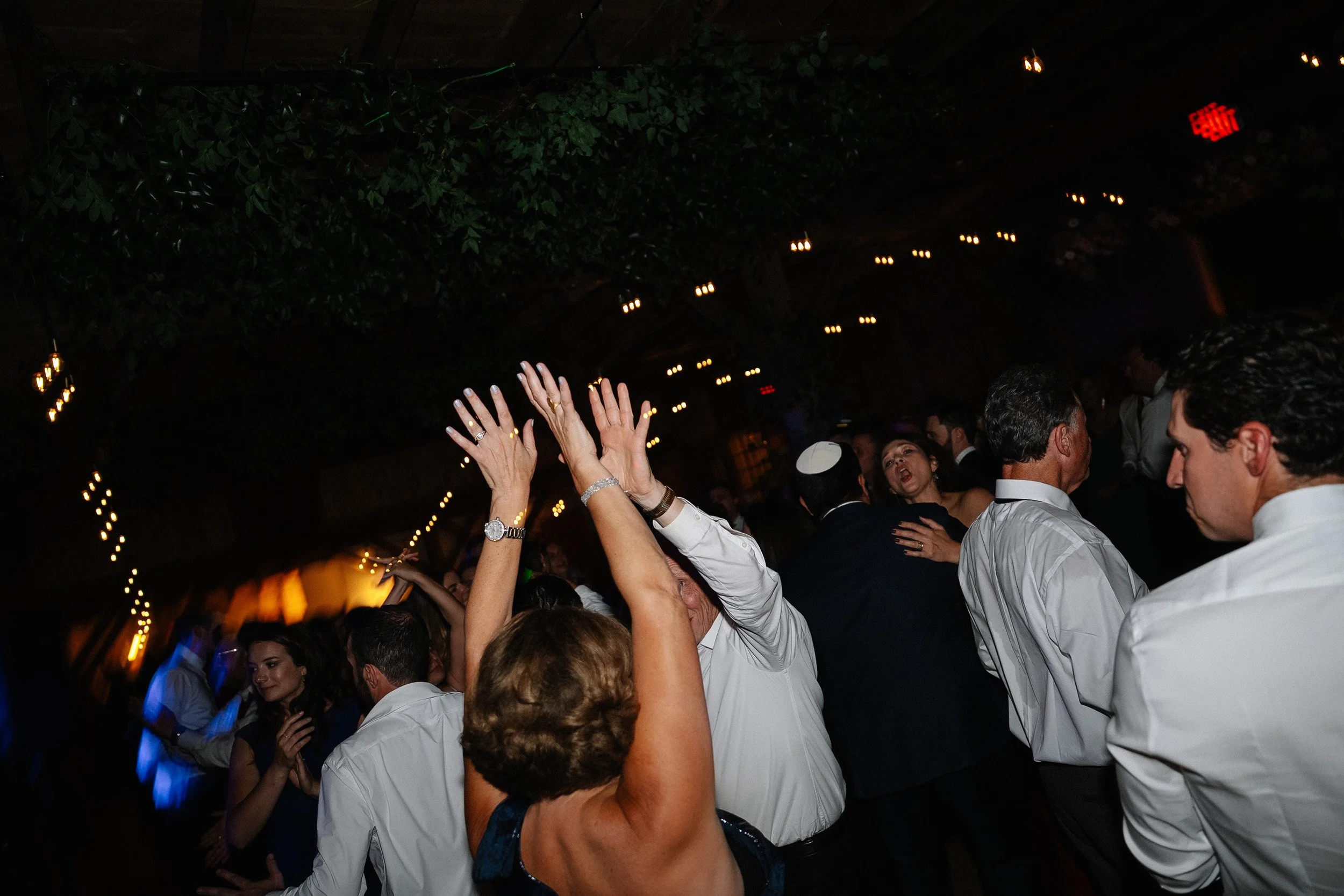People dancing and celebrating at a party or wedding reception, with upper body contact, in a dimly lit venue decorated with string lights and greenery.