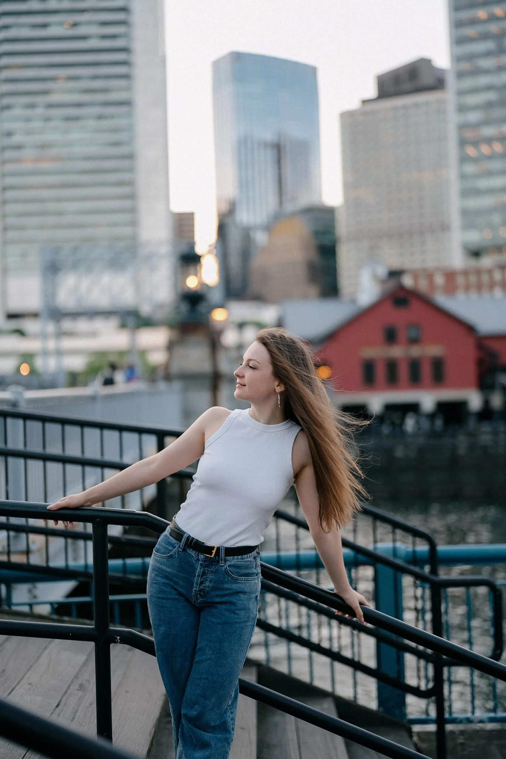 A woman with long hair leaning on a black railing along a waterfront with a cityscape in the background. She is wearing a sleeveless white top and blue jeans, with her eyes closed and a peaceful expression.