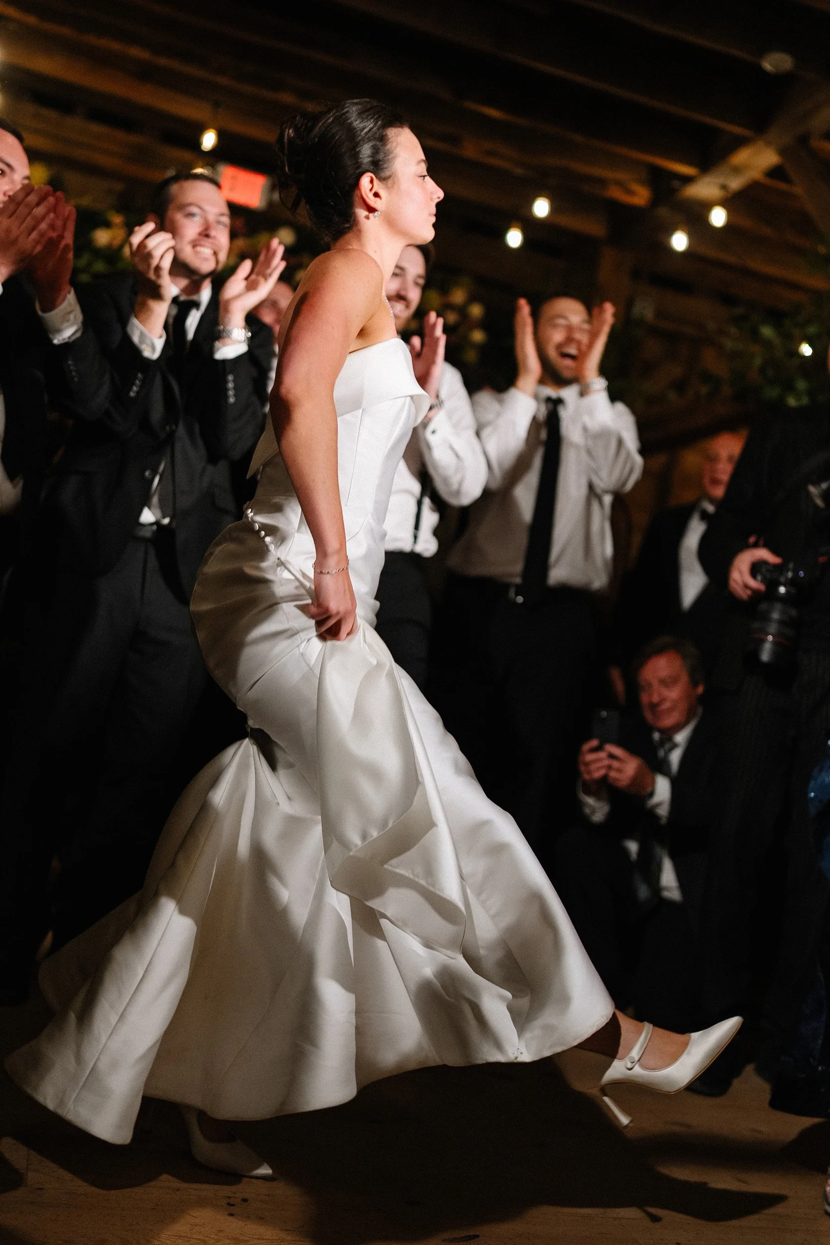 A woman in a white wedding dress is dancing at her wedding reception. She is surrounded by men in suits who are clapping and smiling.