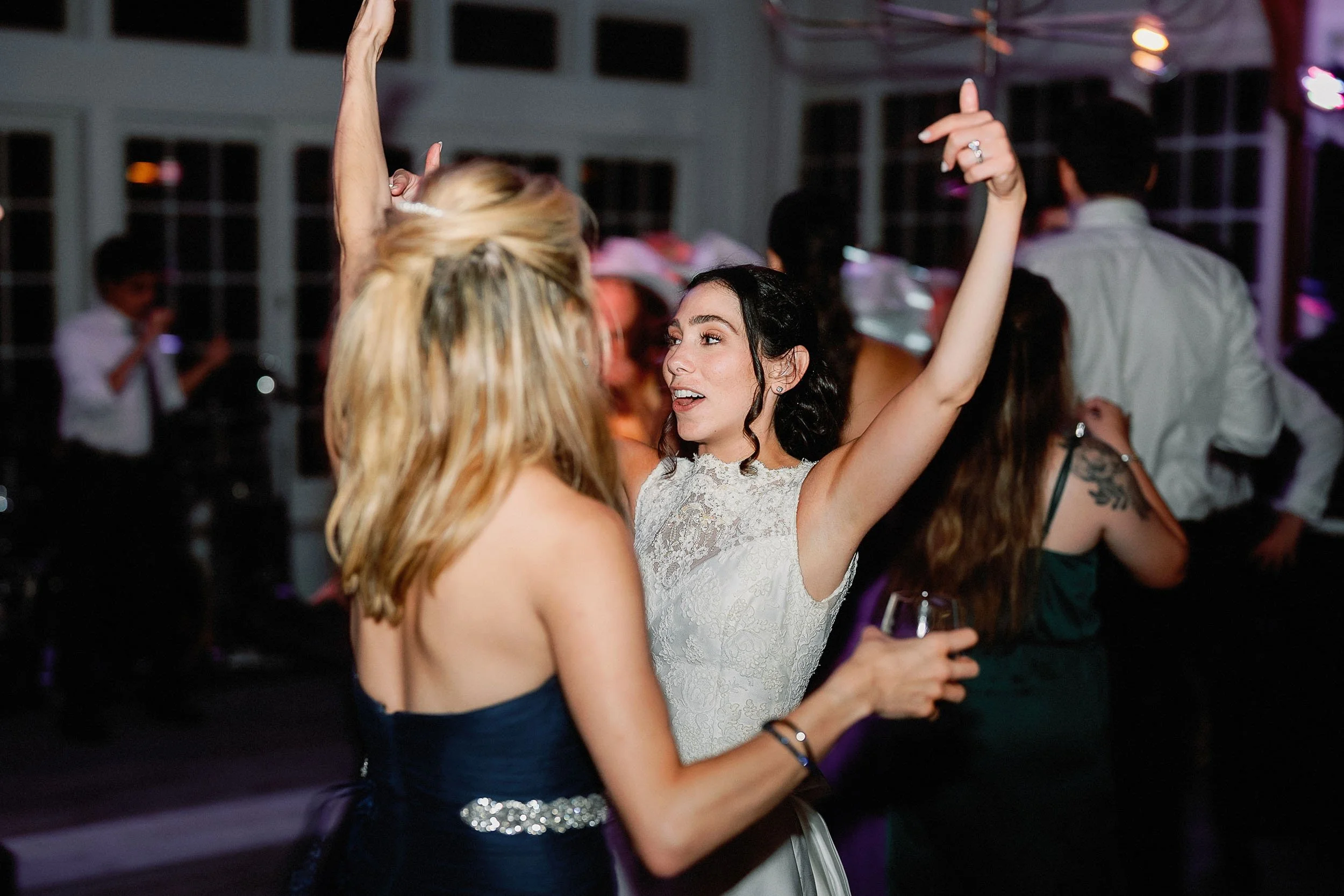 Two women dancing at a party or wedding reception, with one woman in a white dress and the other in a dark dress holding a glass of wine.
