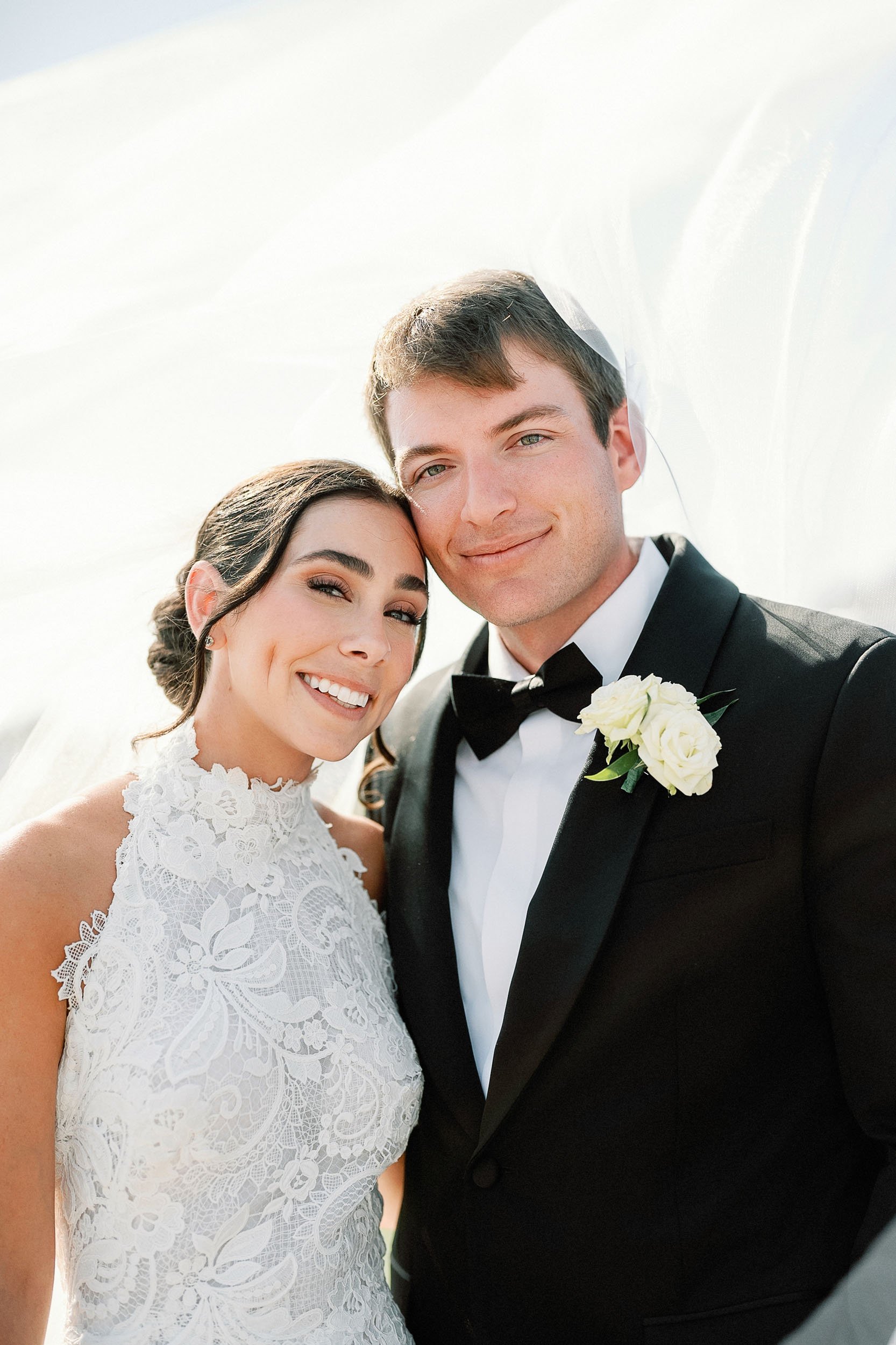 A bride and groom smiling close together outdoors on their wedding day, with the bride in a lace wedding dress and the groom in a black tuxedo with a white shirt and black bow tie.