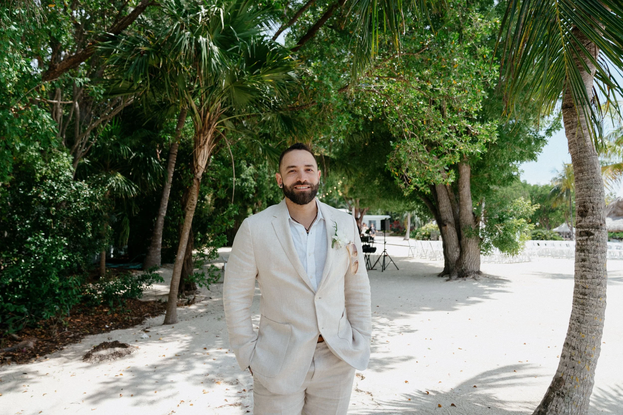 A man in a light-colored suit standing on a sandy beach with tropical trees and greenery in the background, smiling.