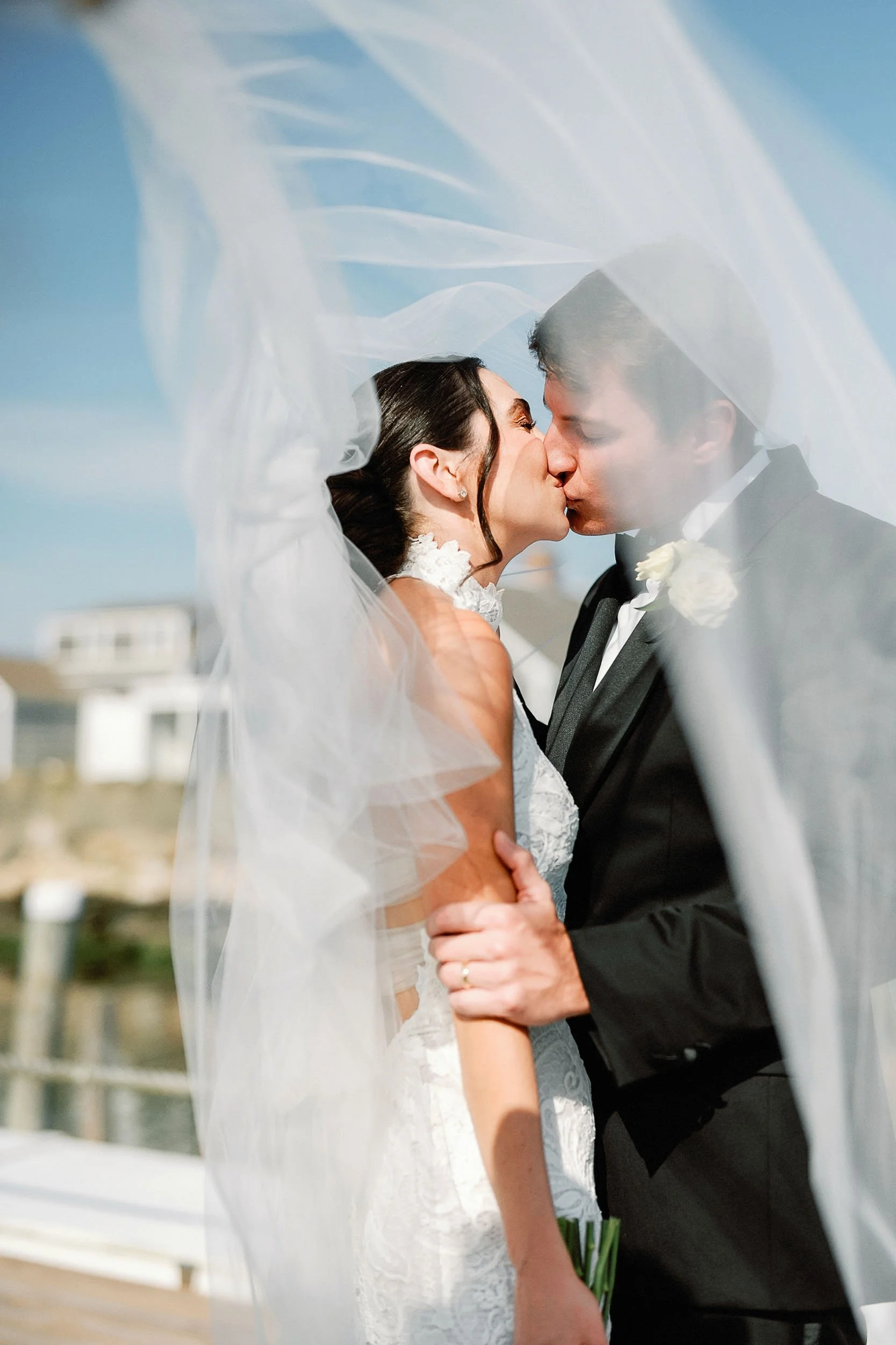 A couple in wedding attire sharing a kiss under a flowing veil on a sunny outdoor day, with a blue sky and beach houses in the background.