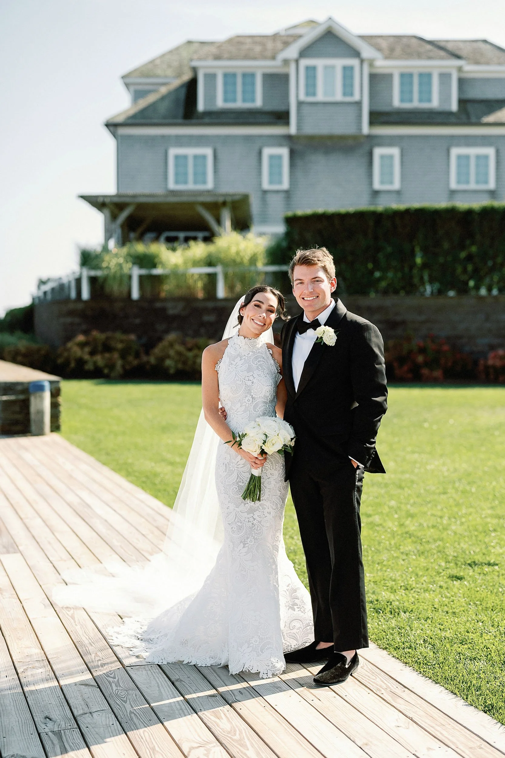 A bride and groom smile and pose together outdoors on a wooden deck, with the bride holding a bouquet of white roses and wearing a lace wedding dress with a veil, and the groom dressed in a black tuxedo with a bowtie, in front of a large house with a