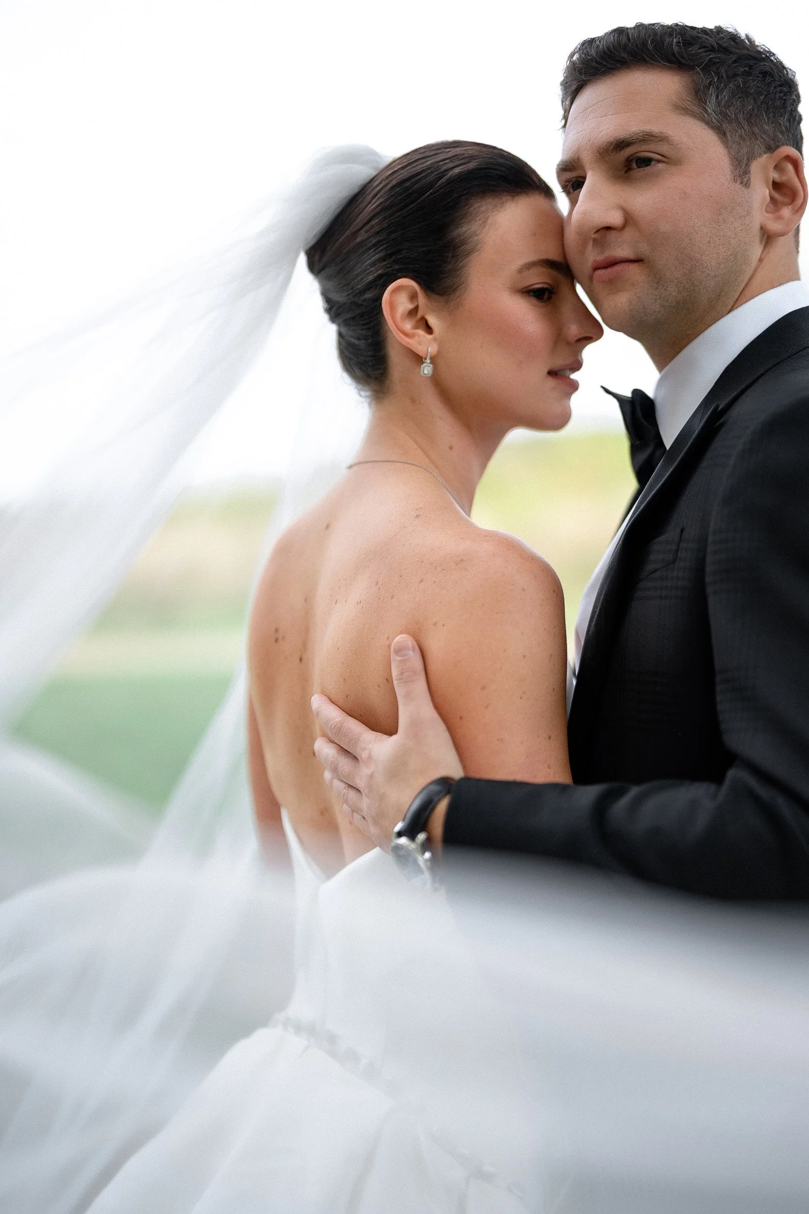 A bride and groom embrace outdoors, with the bride in a strapless dress and veil, and the groom in a tuxedo, both gazing into the distance.