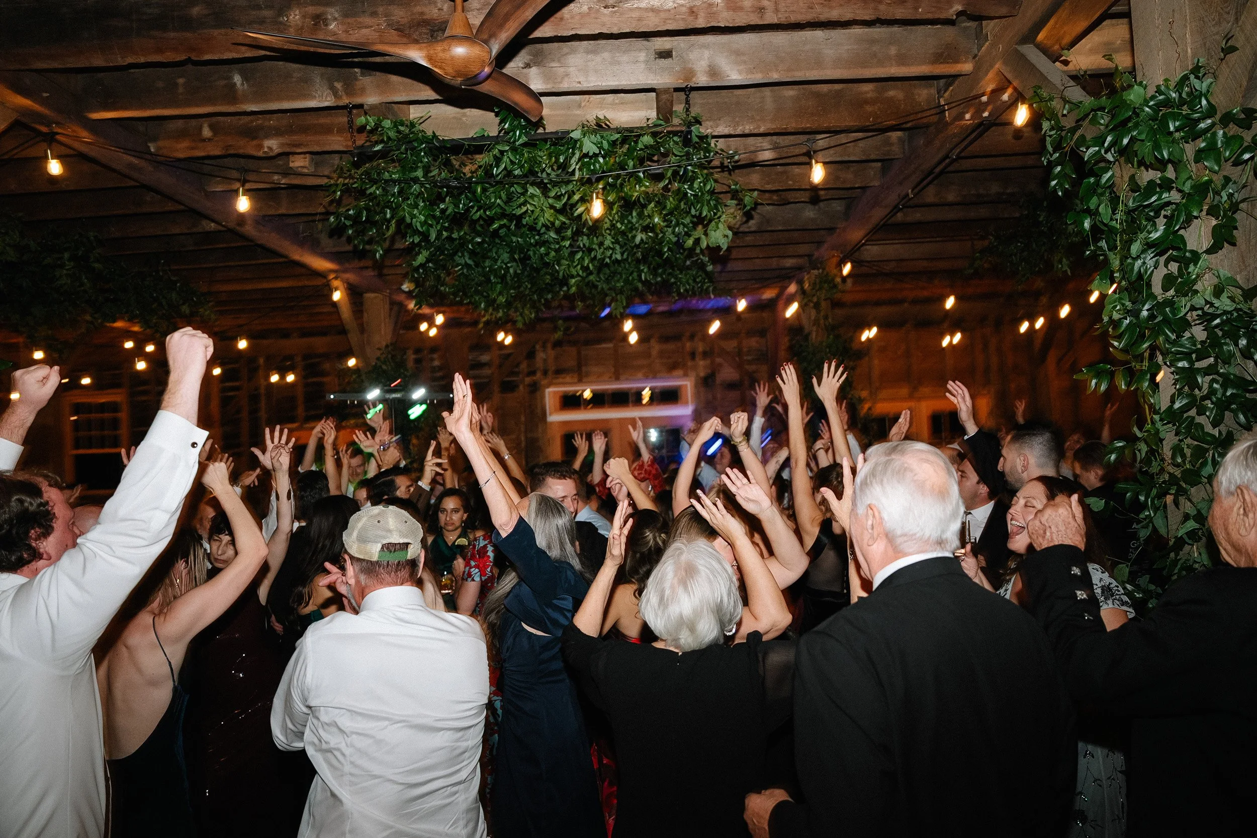 People dancing and celebrating at a party in a rustic barn with hanging lights and greenery.