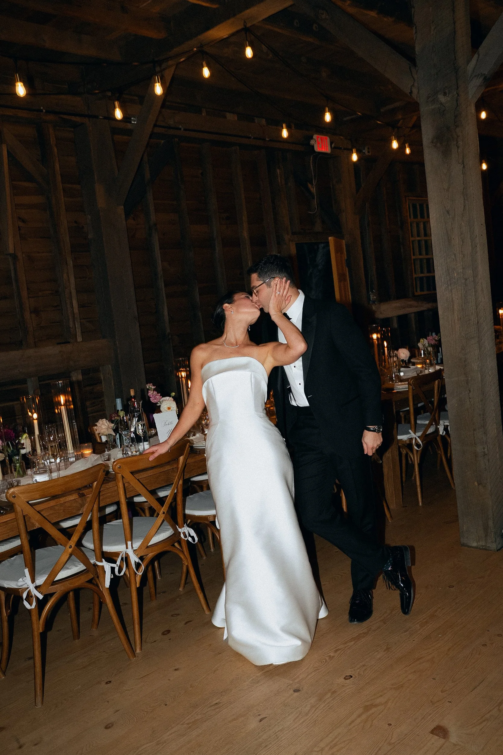 Bride and groom sharing a kiss at their wedding reception in a rustic wooden venue with string lights.