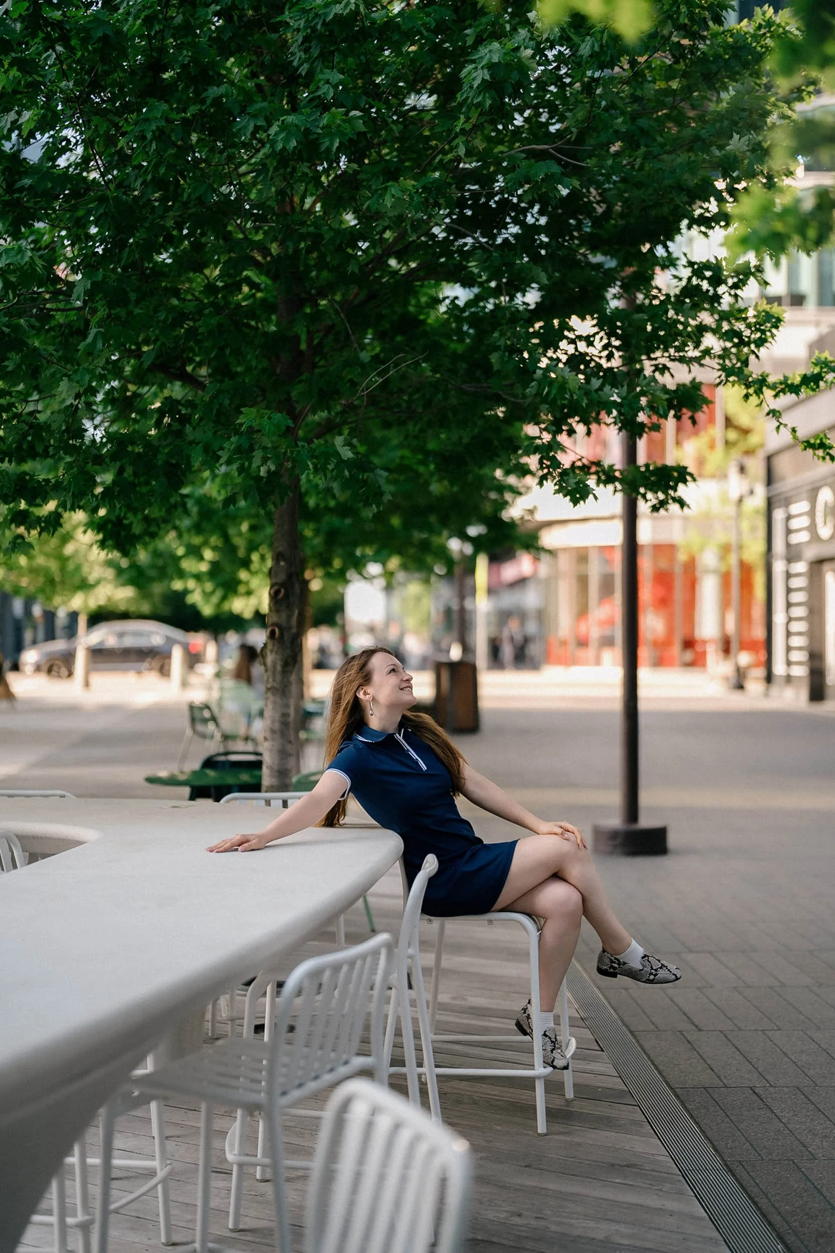 A woman with long hair, wearing a navy blue dress and snake print shoes, sitting on a chair at an outdoor cafe table, smiling and looking up at a tree in an urban setting.