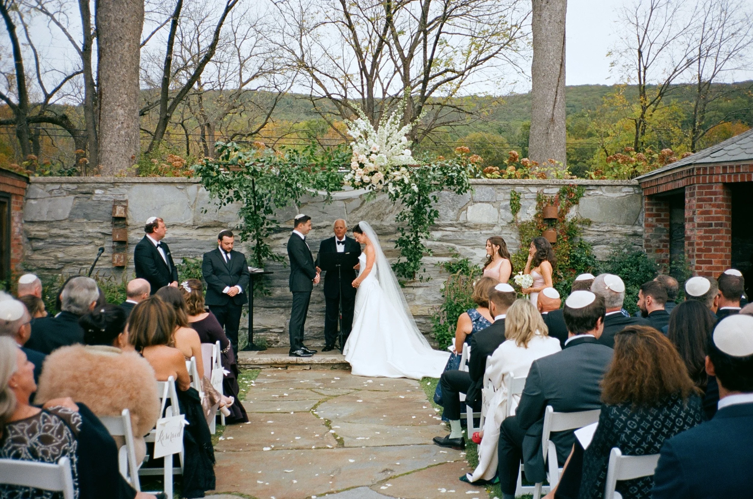 Outdoor wedding ceremony with a bride and groom exchanging vows, surrounded by guests seated in white chairs, overlooking a stone wall decorated with flowers and greenery, with trees and hills in the background.