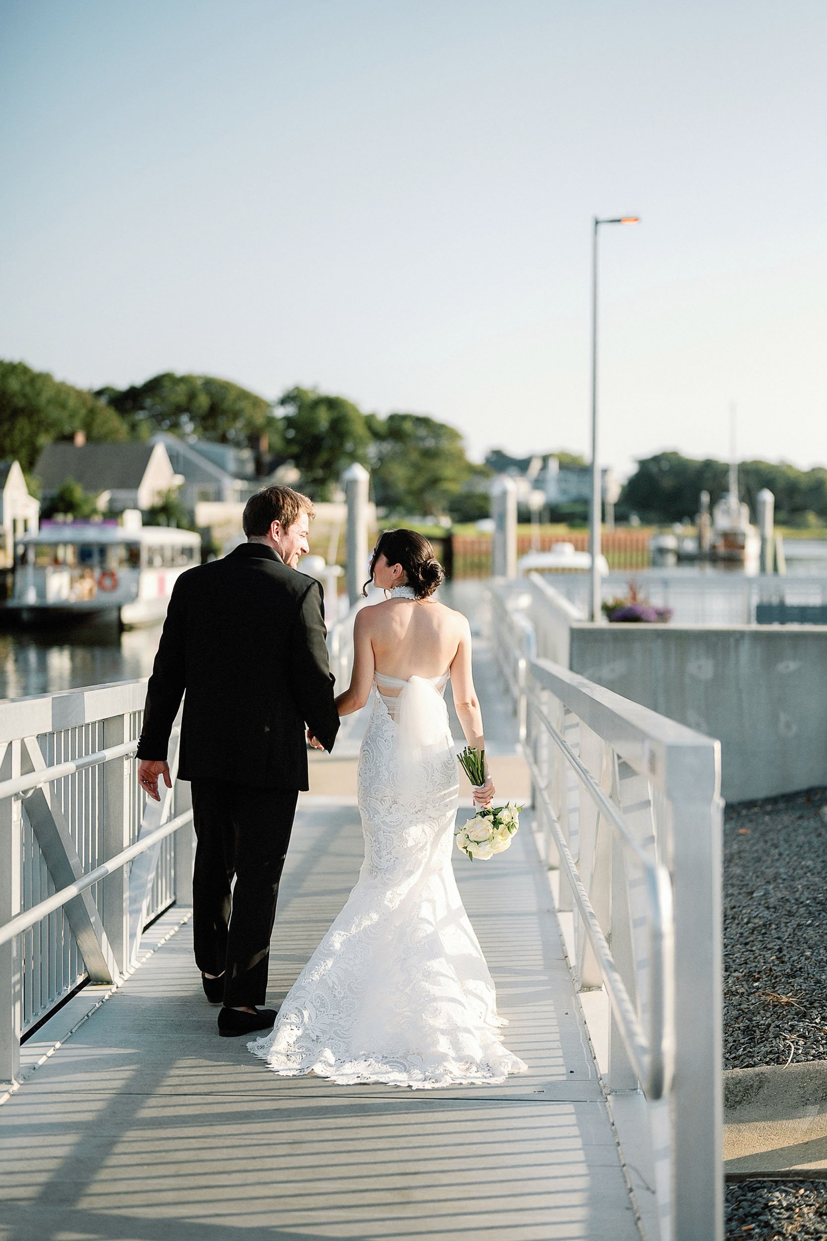 A bride and groom walk arm in arm on a bridge near the water, with the bride holding a bouquet of white flowers, during sunset.