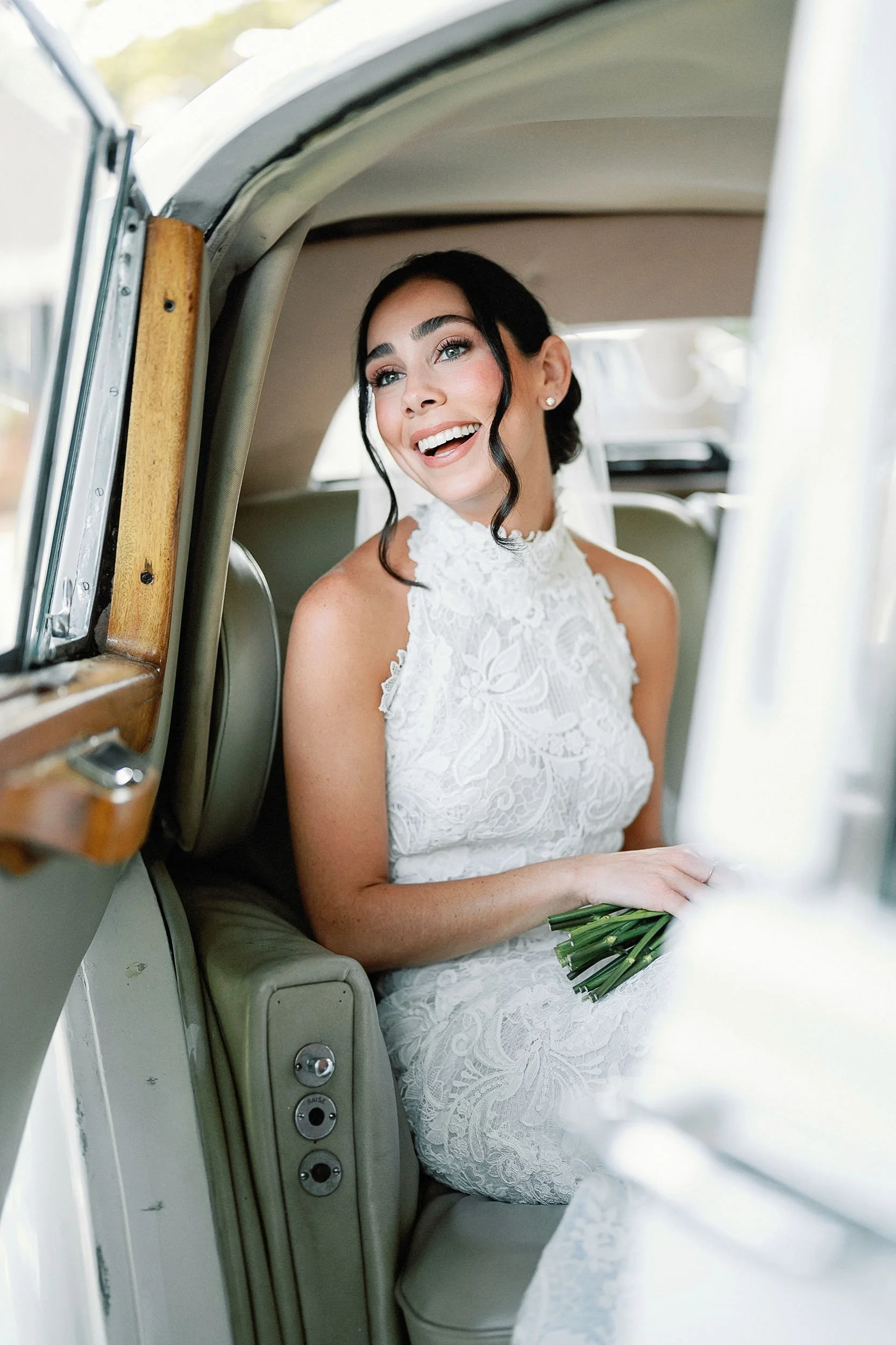 A bride in a white lace dress smiling while sitting inside a vintage car, holding a small bouquet of green flowers.