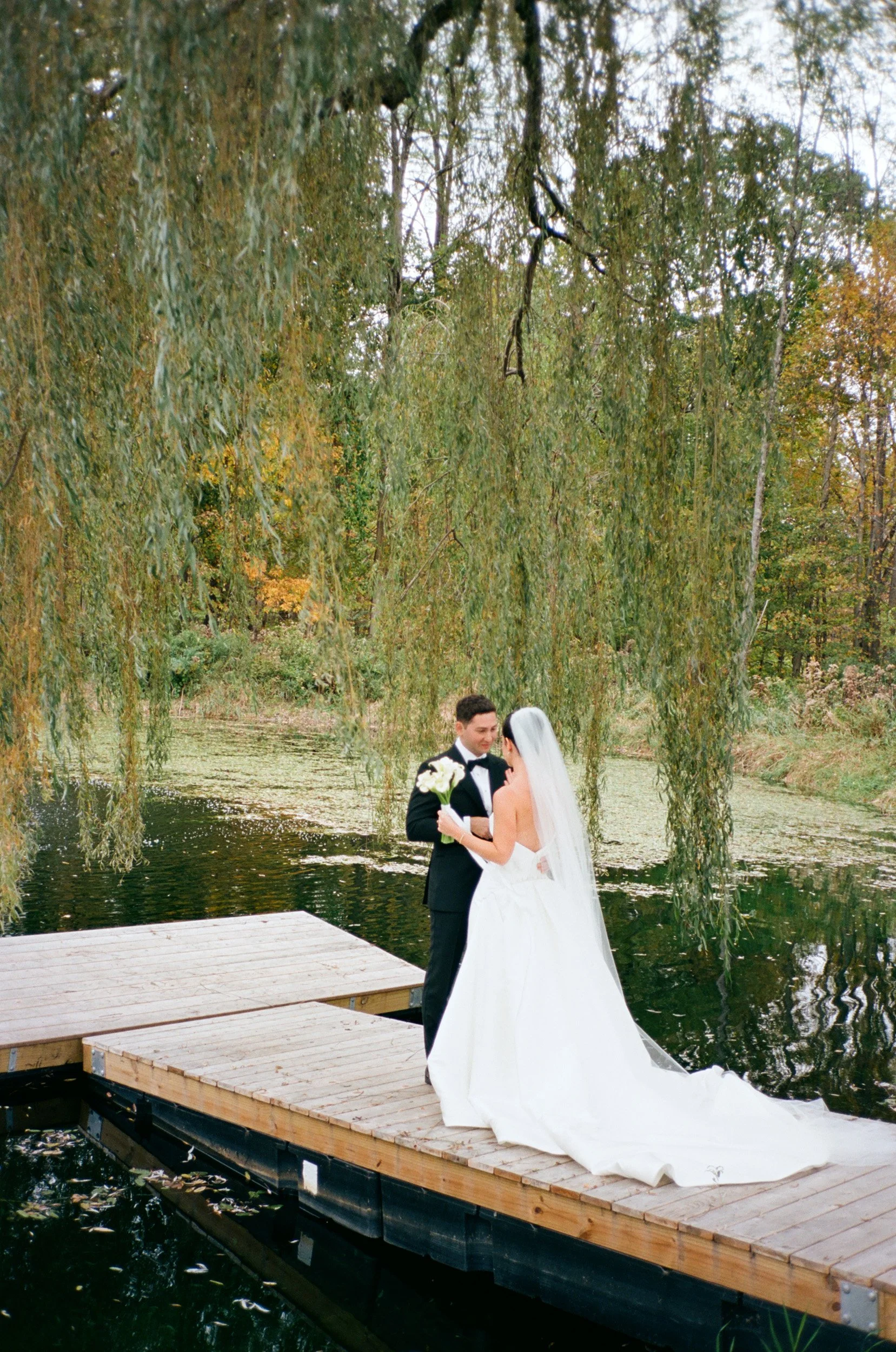 Bride and groom standing on a wooden dock by a lake, surrounded by trees, with the bride holding a bouquet of white flowers.