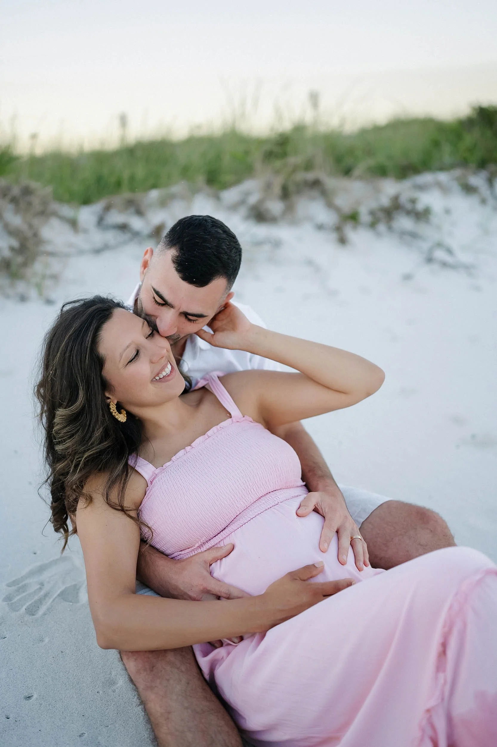 A couple sitting on the sand at the beach, with the woman pregnant, sharing a tender moment and smiling.