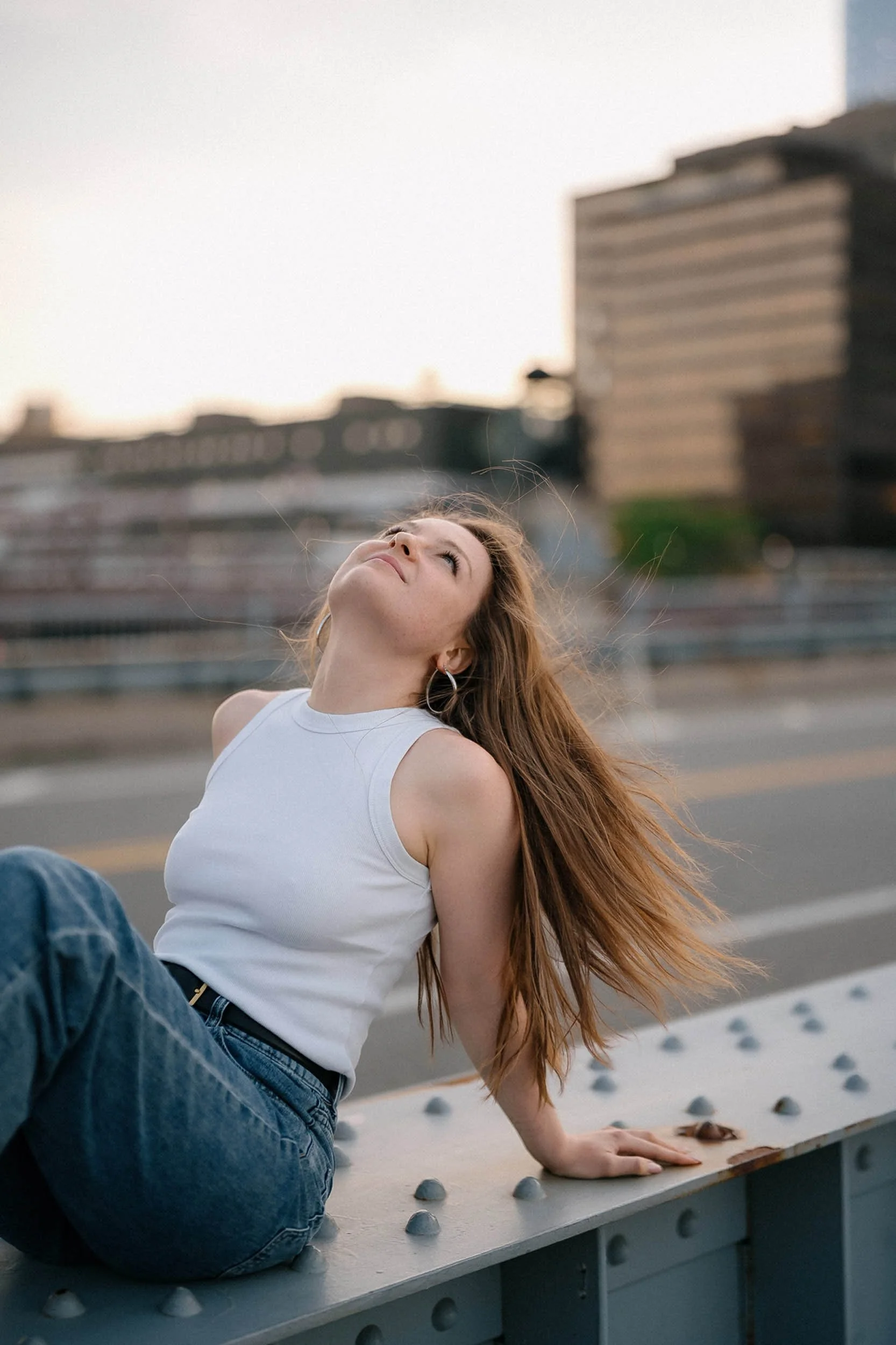 A young woman with long red hair sitting on a bridge railing in an urban setting, looking upward with a relaxed and peaceful expression.