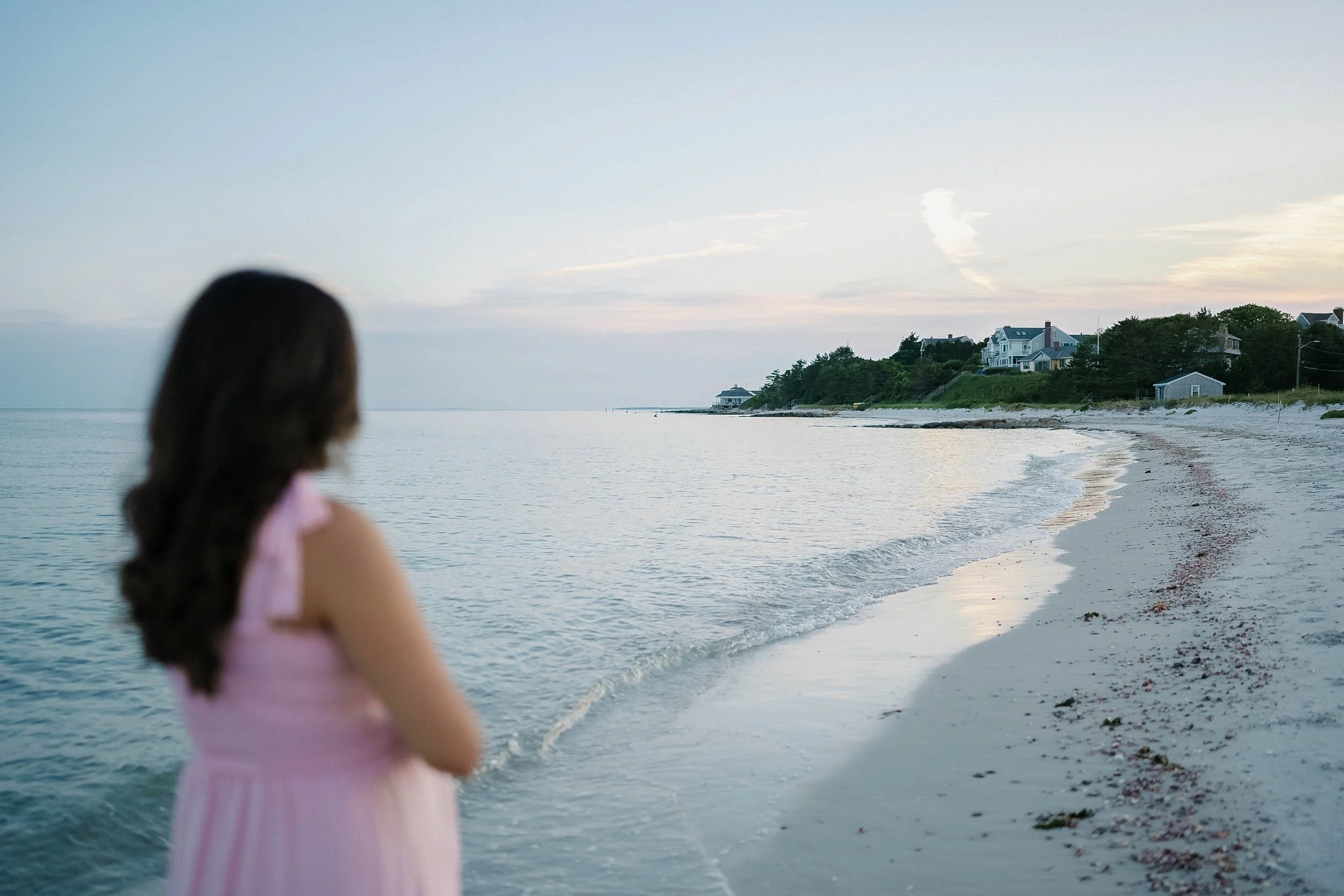 A woman with long dark hair standing on a sandy beach looking out at the ocean during sunset or dawn, with houses and trees on the shoreline in the distance.