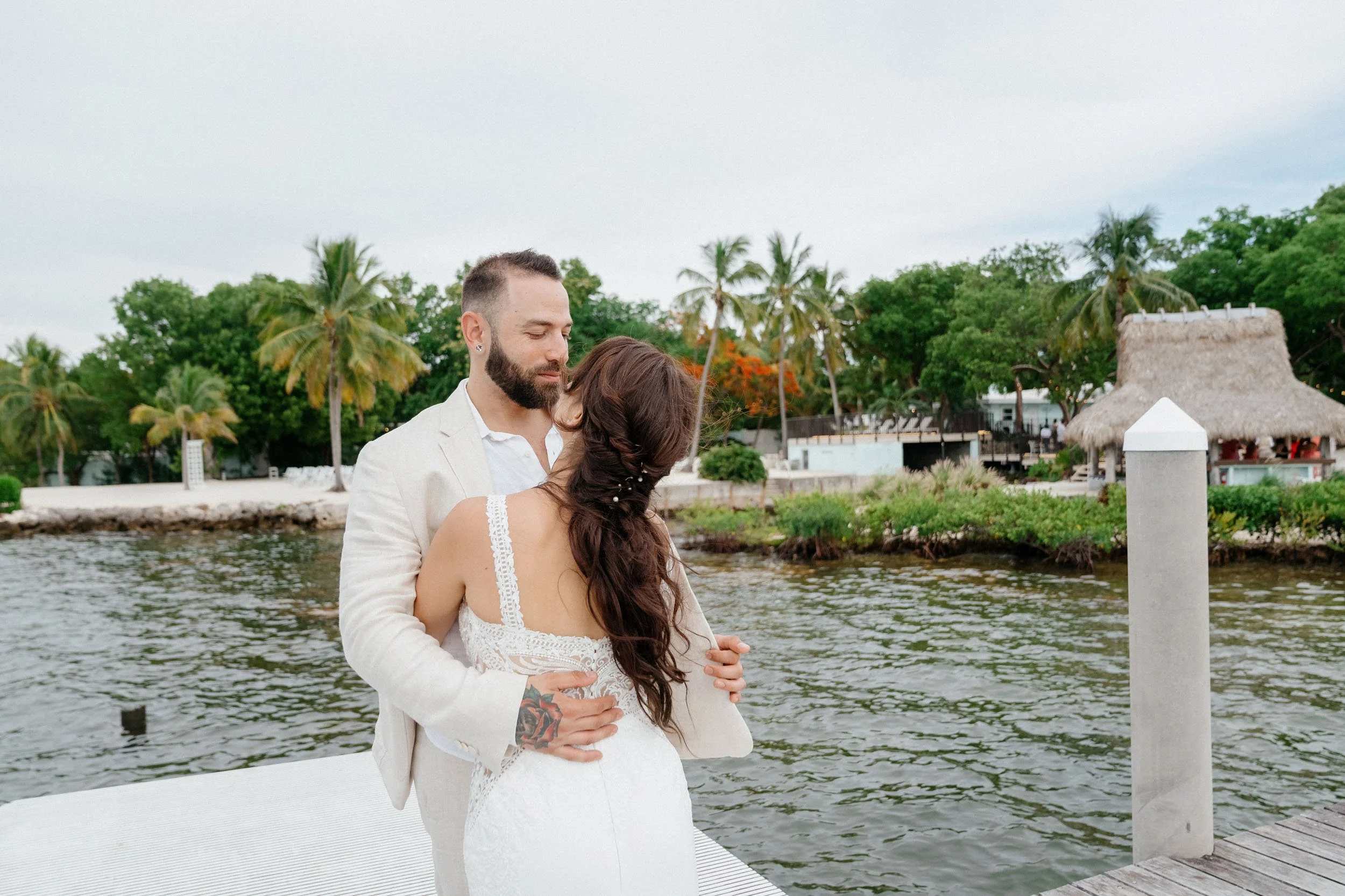 A couple embracing by the water with palm trees and a thatched roof hut in the background