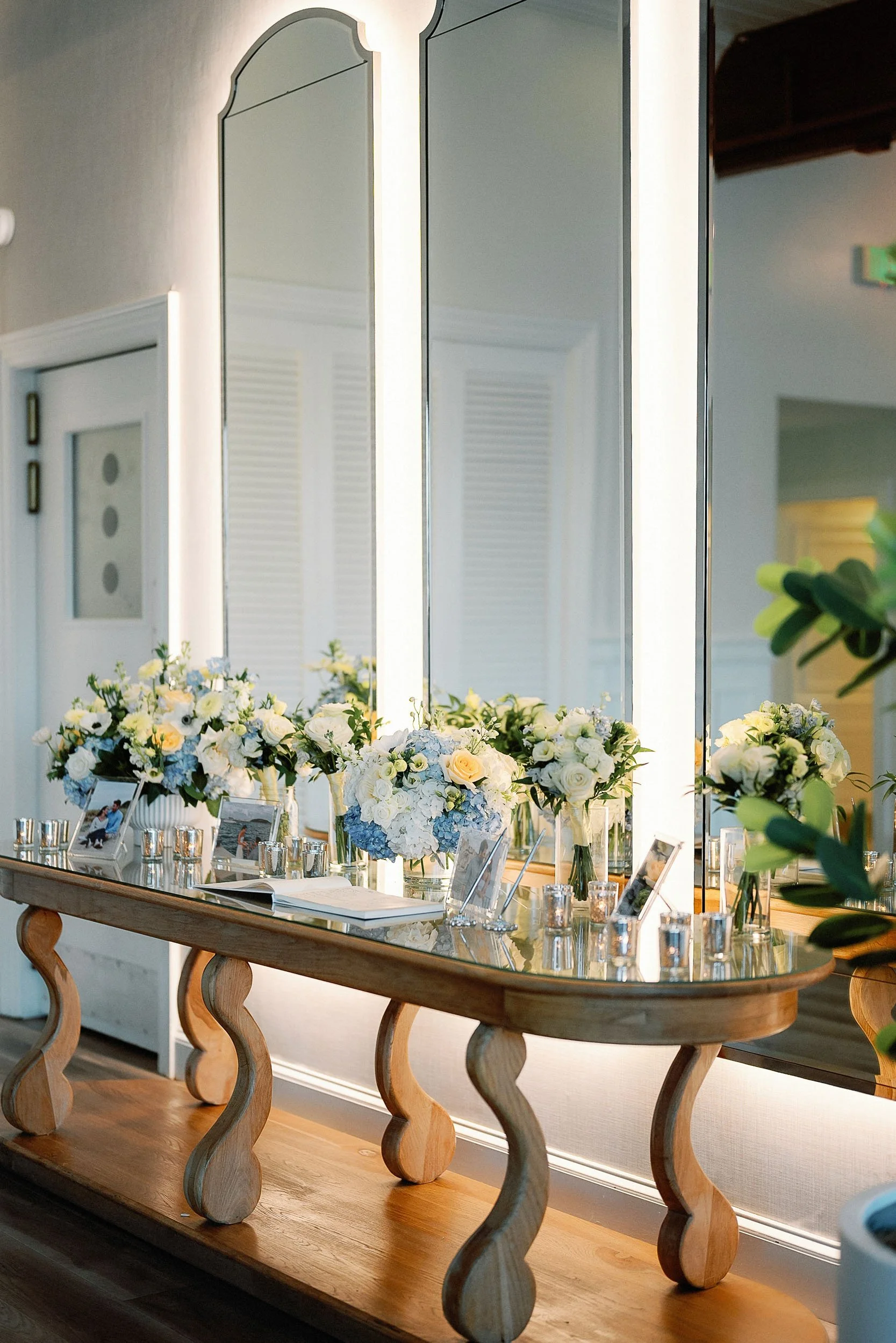 A wooden table decorated with white and light blue flower bouquets, framed photographs, and glass candle holders, placed in front of large illuminated mirrors.