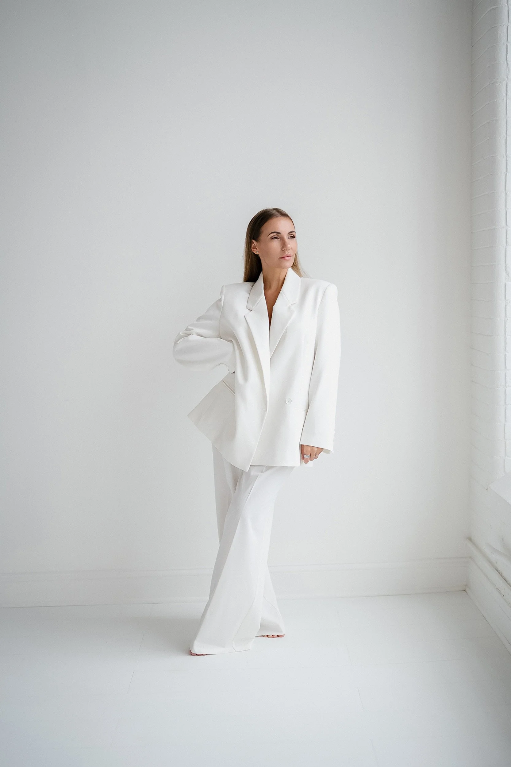 A woman in a white suit standing in a bright, minimalistic room with a white brick wall and large window.