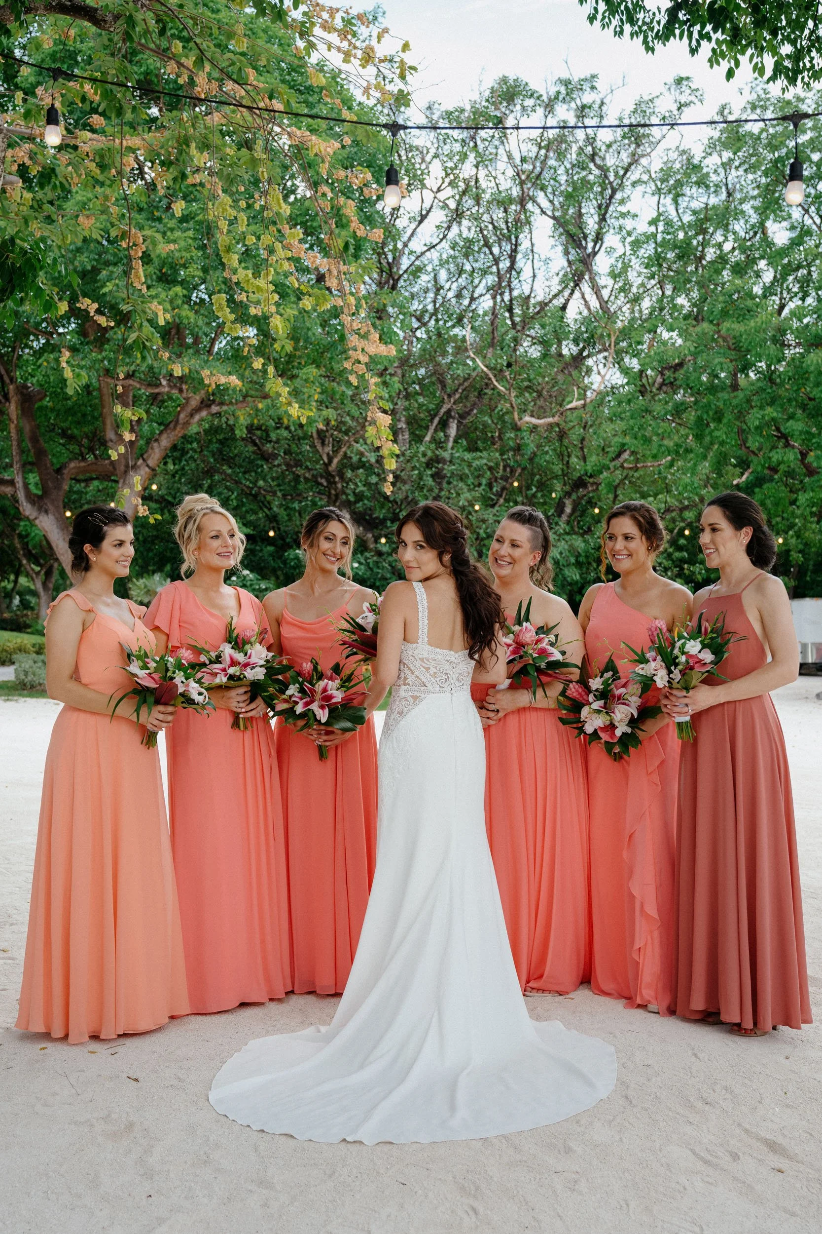 Bride in a white wedding dress with lace details posing with six bridesmaids in coral dresses, holding bouquets of pink and white flowers, outdoors beneath string lights and surrounded by green trees.