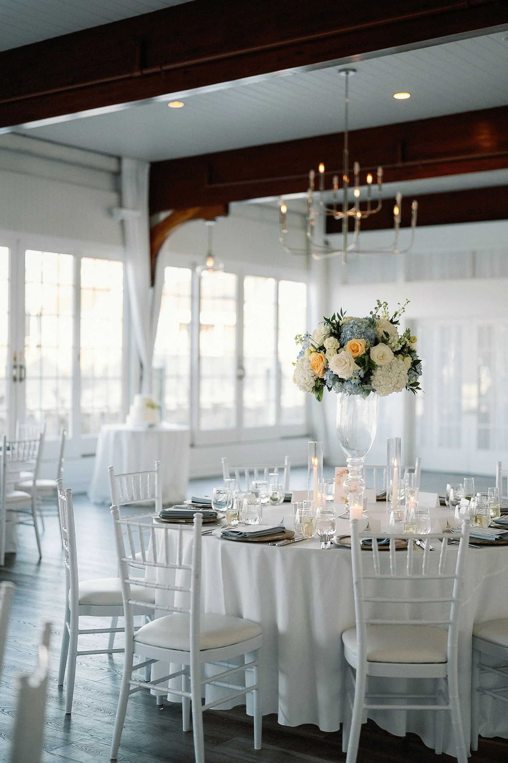 Elegant banquet table setup with white tablecloth, floral centerpiece, candles, glassware, and dishes, in a bright room with large windows and a chandelier