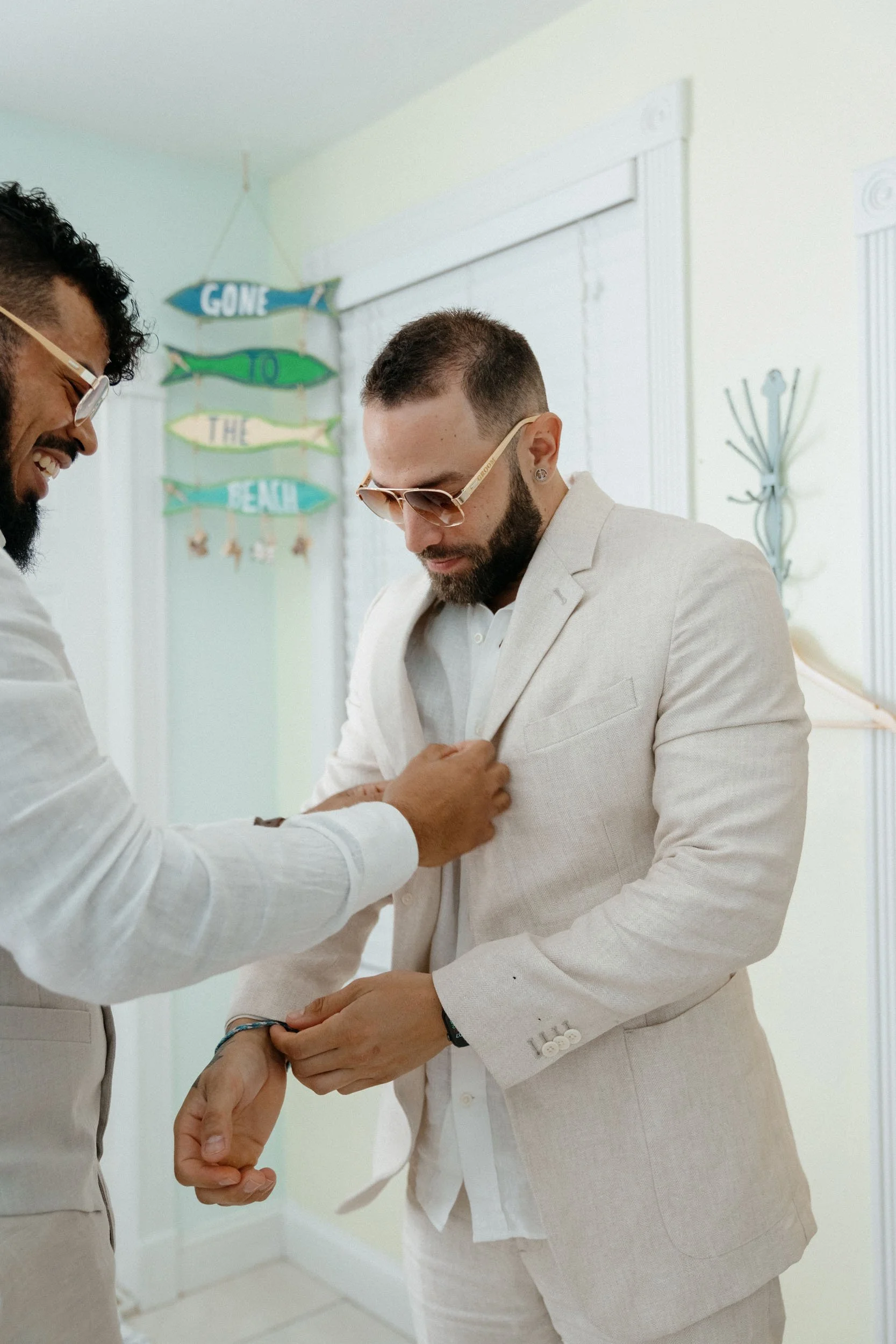 Two men dressed in light-colored suits, one adjusting the other's sleeve, inside a bright room with seaside-themed wall decorations.