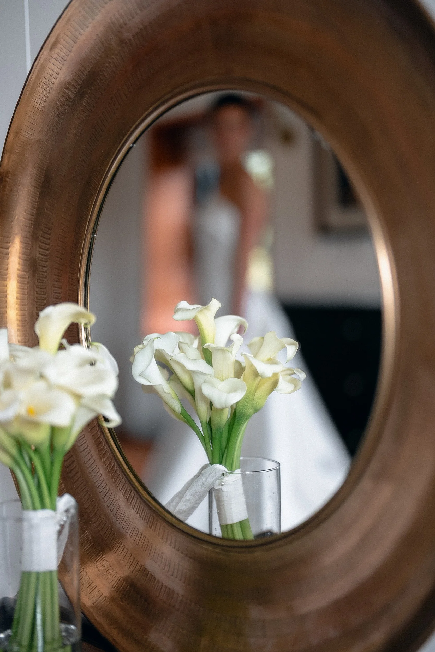A mirror reflects a woman in a wedding dress holding a bouquet, with white calla lilies in a clear vase in the foreground.