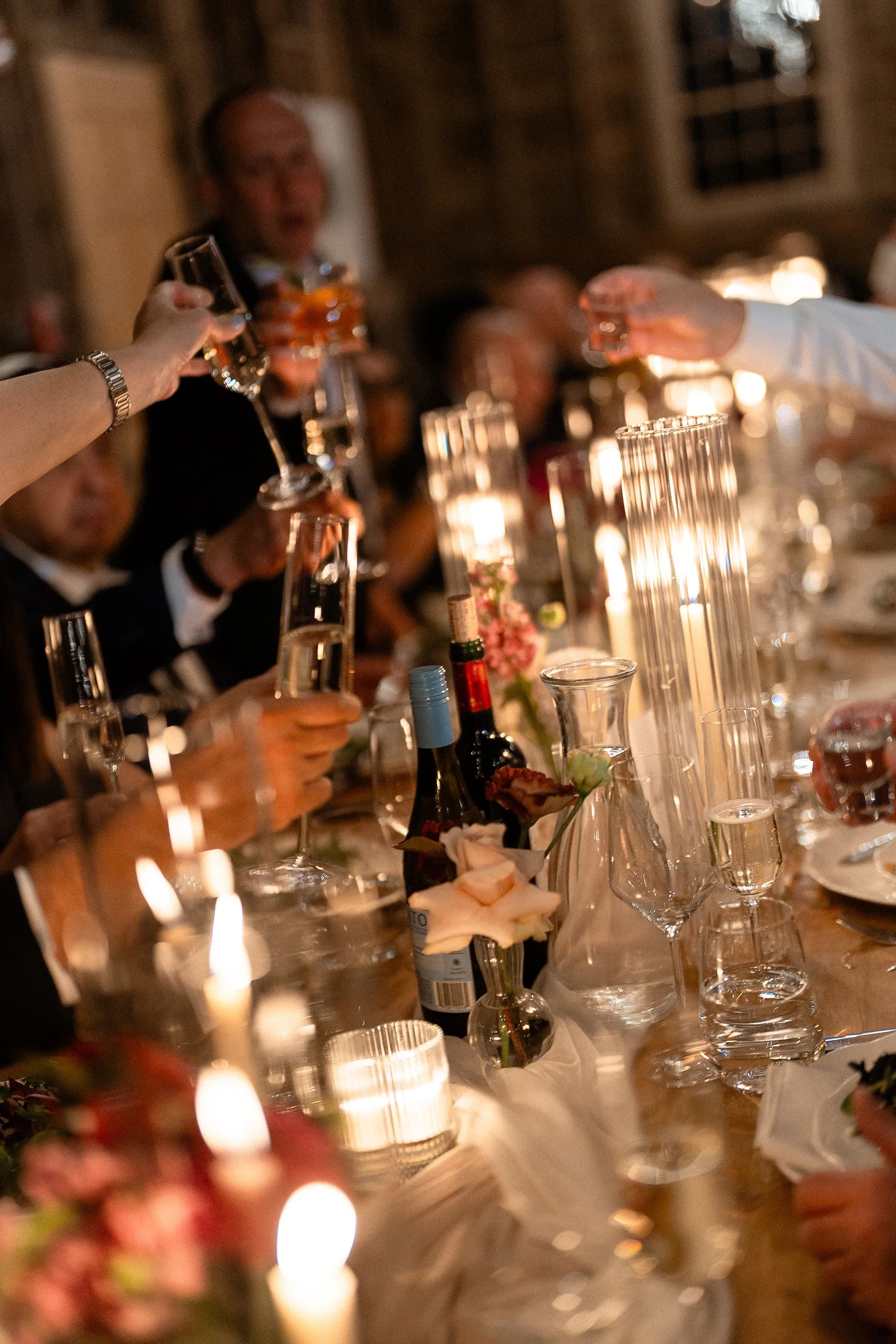 People raising glasses in a toast at a dinner party with candles and wine bottles on a long table.