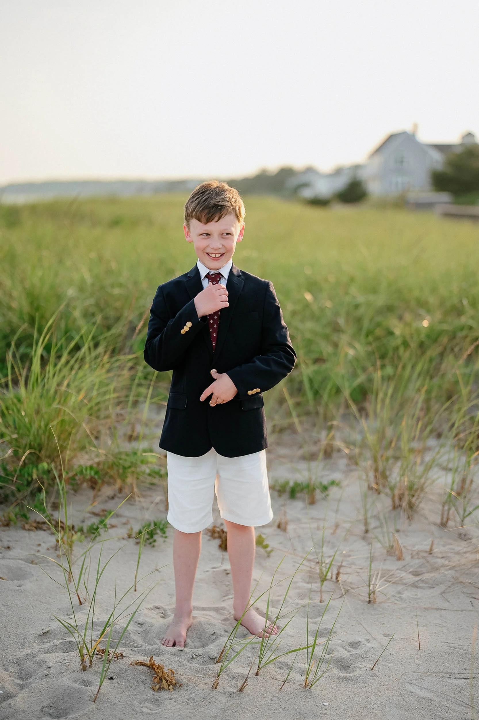 A young boy dressed in a black blazer, white shorts, and a red tie standing barefoot on the sandy ground of a beach, smiling and adjusting his tie with a relaxed background of green grass, houses, and an overcast sky.