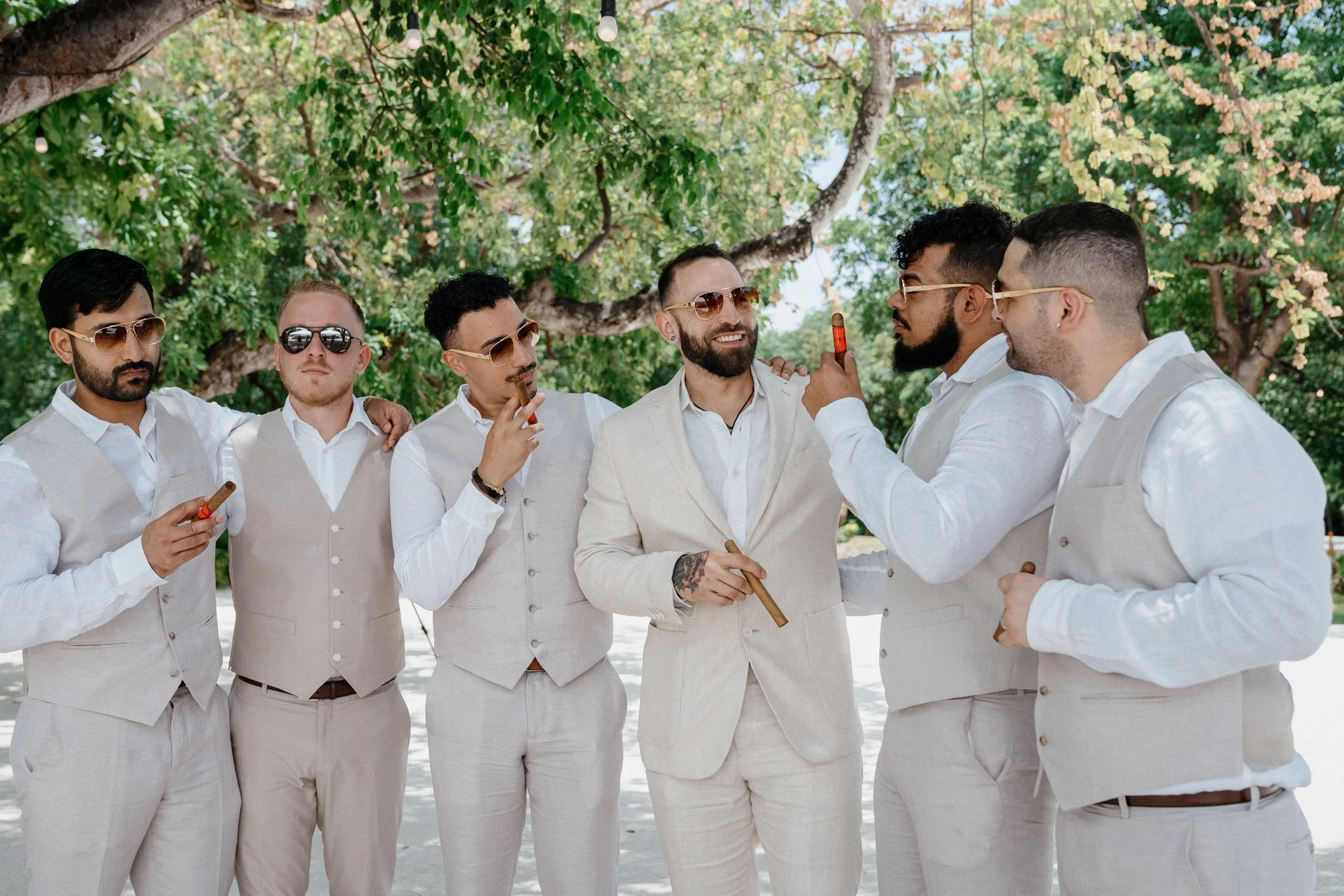 Group of seven men dressed in light-colored suits, wearing sunglasses, standing outdoors under a tree, holding cigars, engaged in conversation and smiling.