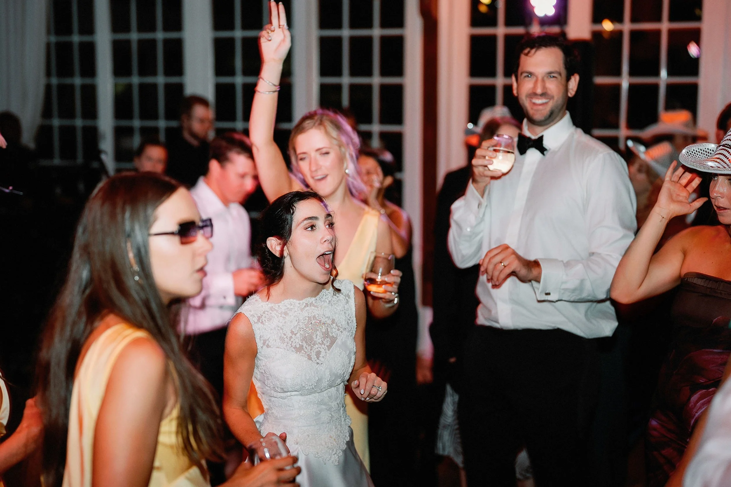 People dancing and socializing at a wedding reception, with a woman in a white wedding dress in the center, raising her hand, and a man in a white shirt and black bow tie holding a drink. The setting is indoors with large windows in the background.