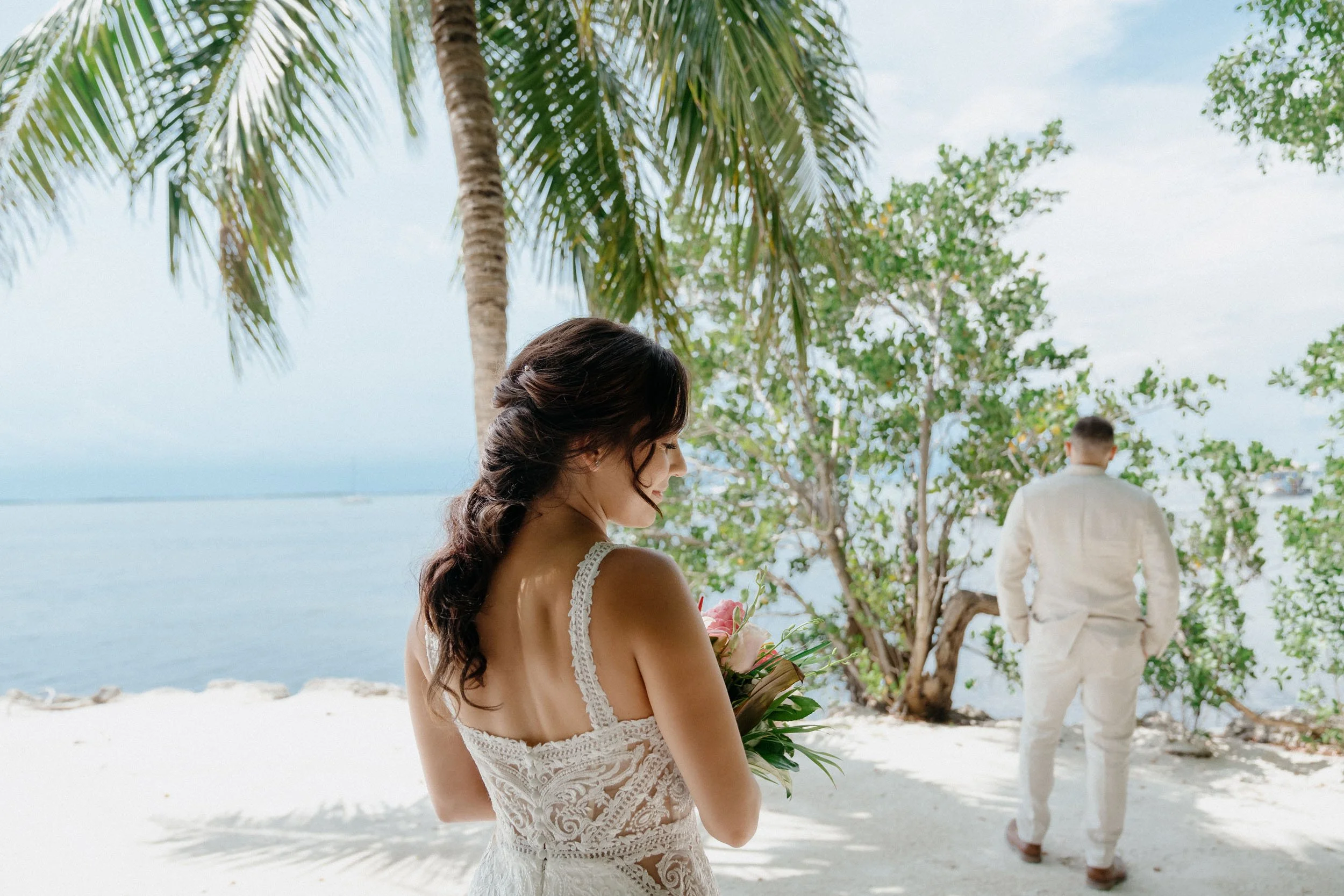 A woman in a lace white dress holding a bouquet of pink flowers on a beach with palm trees, while a man in a white suit walks away in the background.