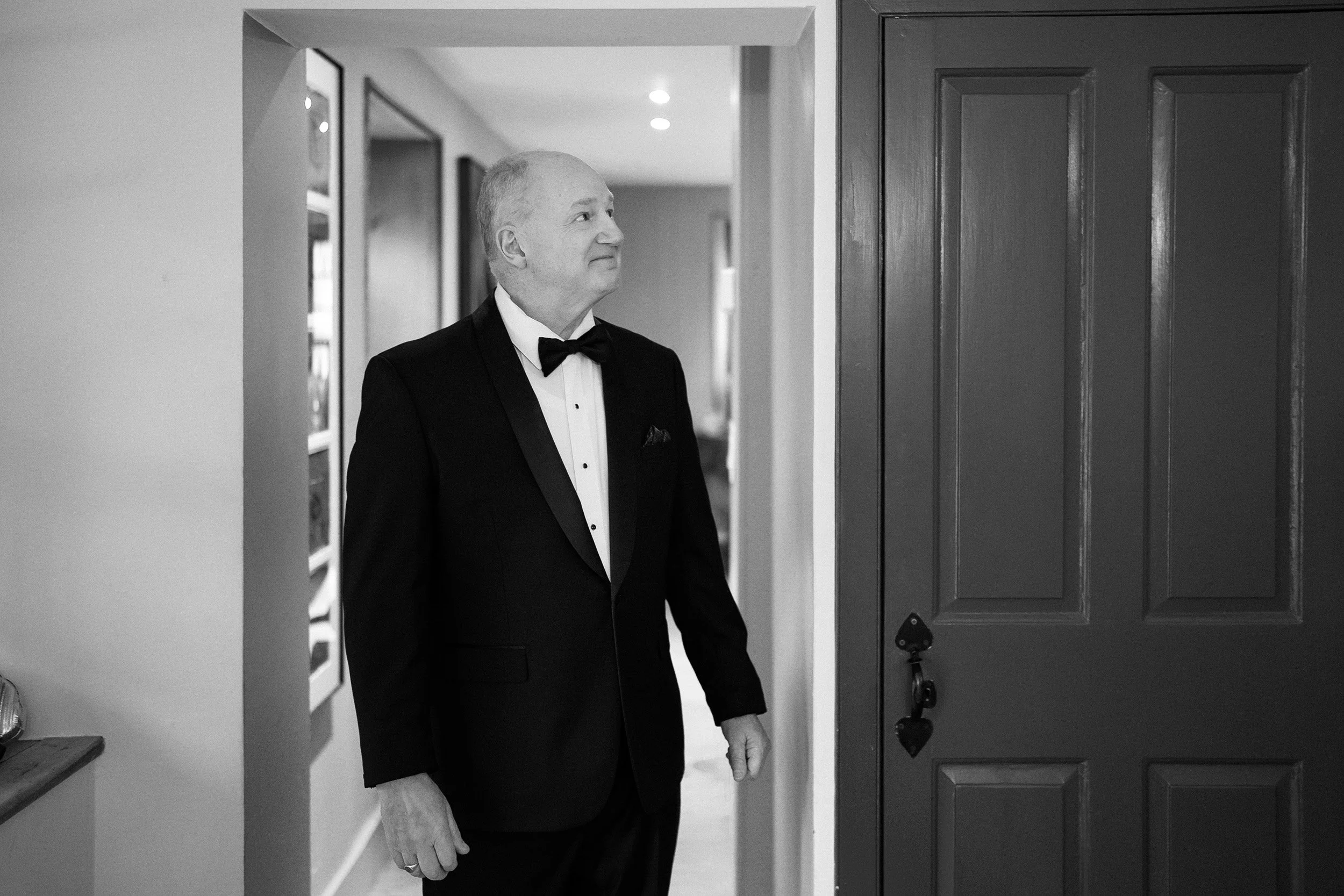 An elderly man in a black tuxedo with a bow tie standing at an open door, looking to his right, in an indoor setting.
