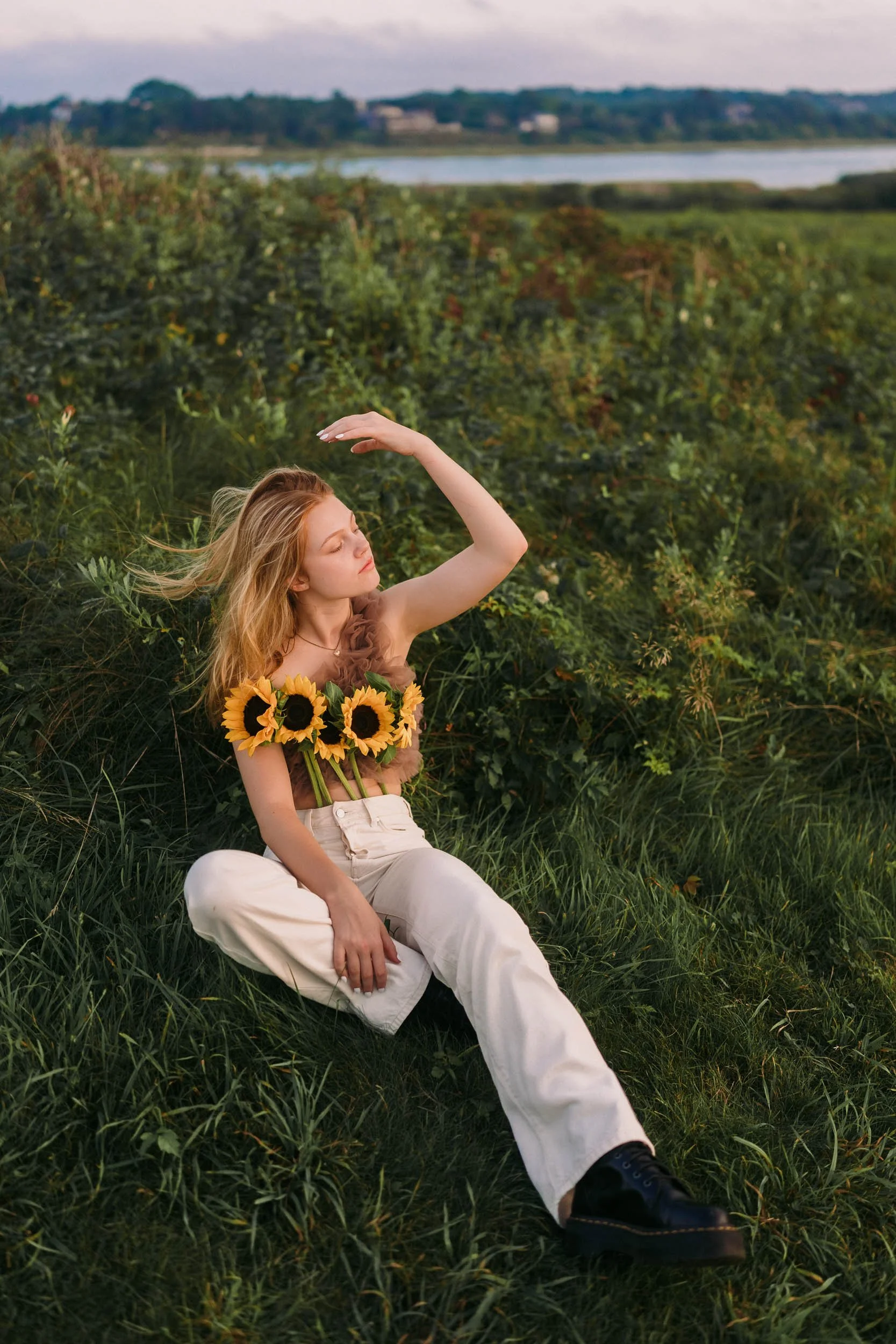 A young woman with long blonde hair sitting on grass in a field, wearing white pants and black shoes, with a floral top decorated with sunflowers, posing with her hand over her eyes, and surrounded by green bushes and distant water and trees.