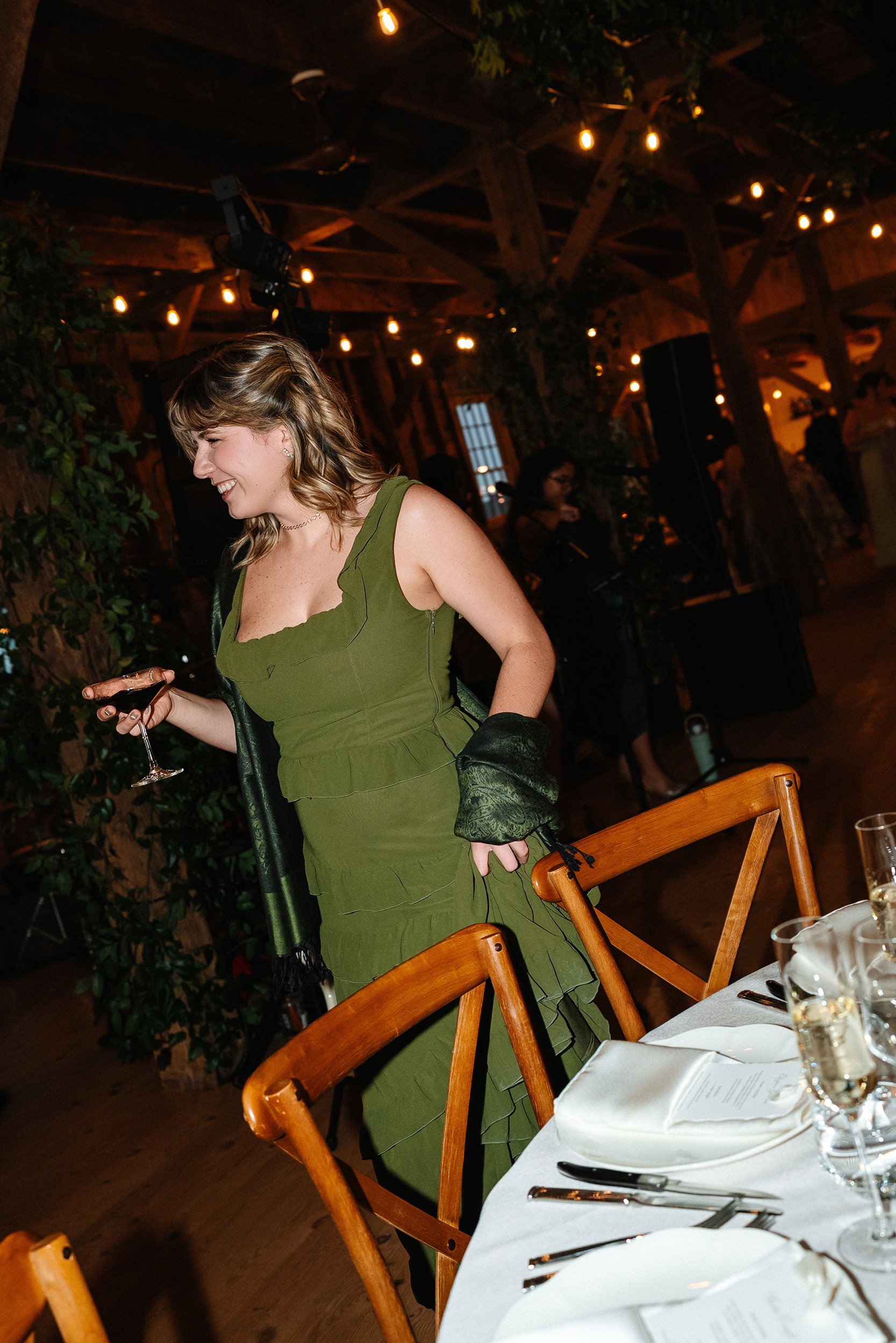 A woman in a green dress holding a glass of red wine standing next to a dining table with place settings, in a warmly lit rustic venue with wooden beams and string lights.