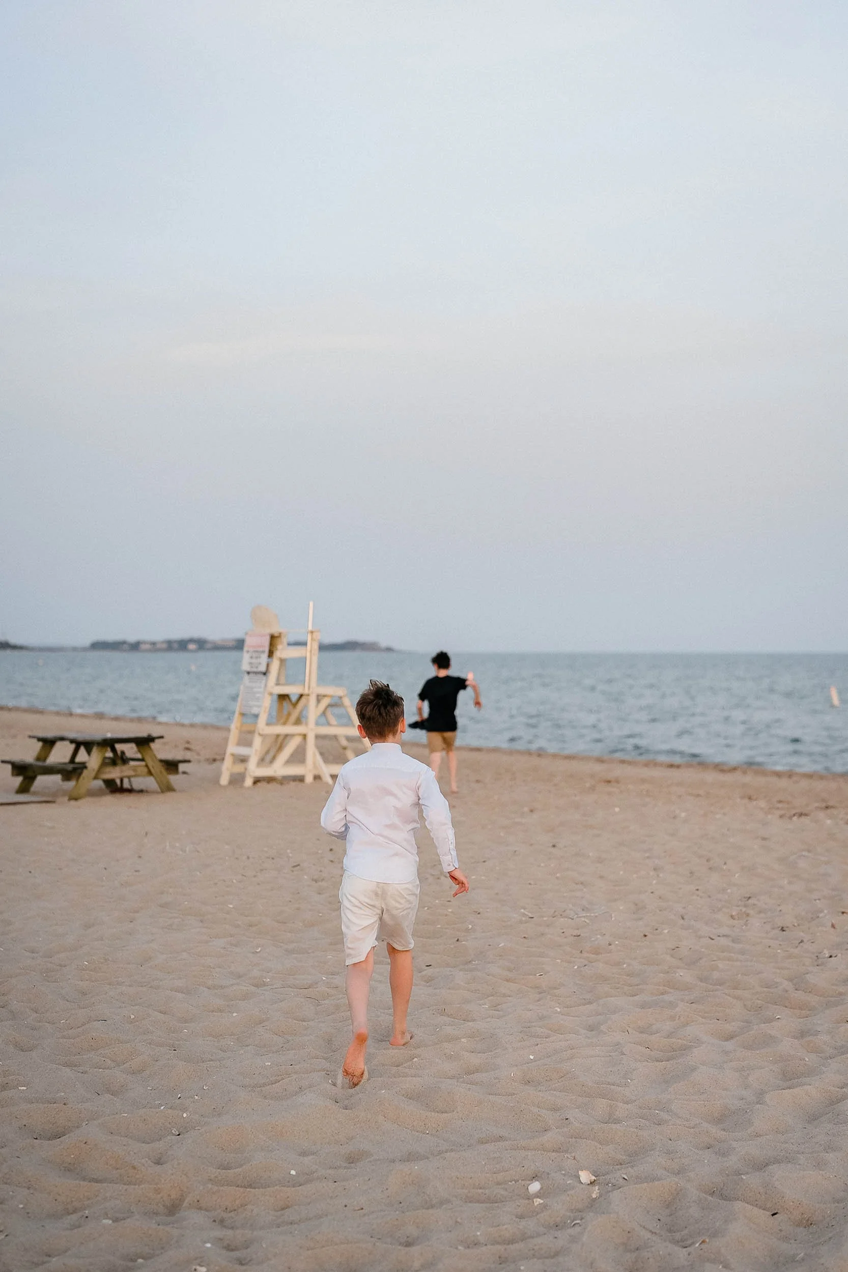 Two boys are running on the sandy beach towards the water, with the shoreline and calm sea in the background.