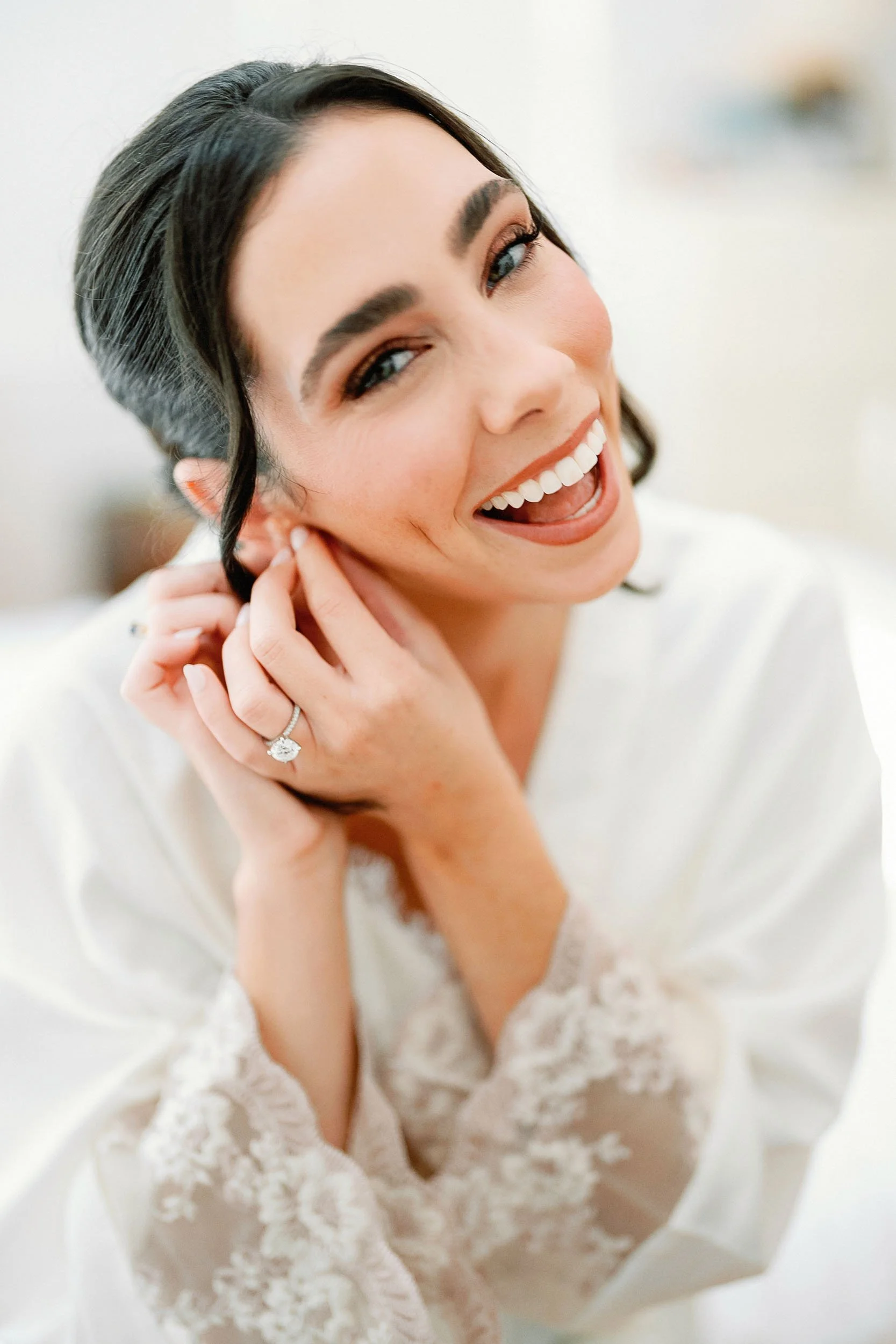 A woman with dark hair, bright smile, and earrings is putting on an earring, wearing a white satin robe with lace cuffs, and a diamond ring on her left hand.