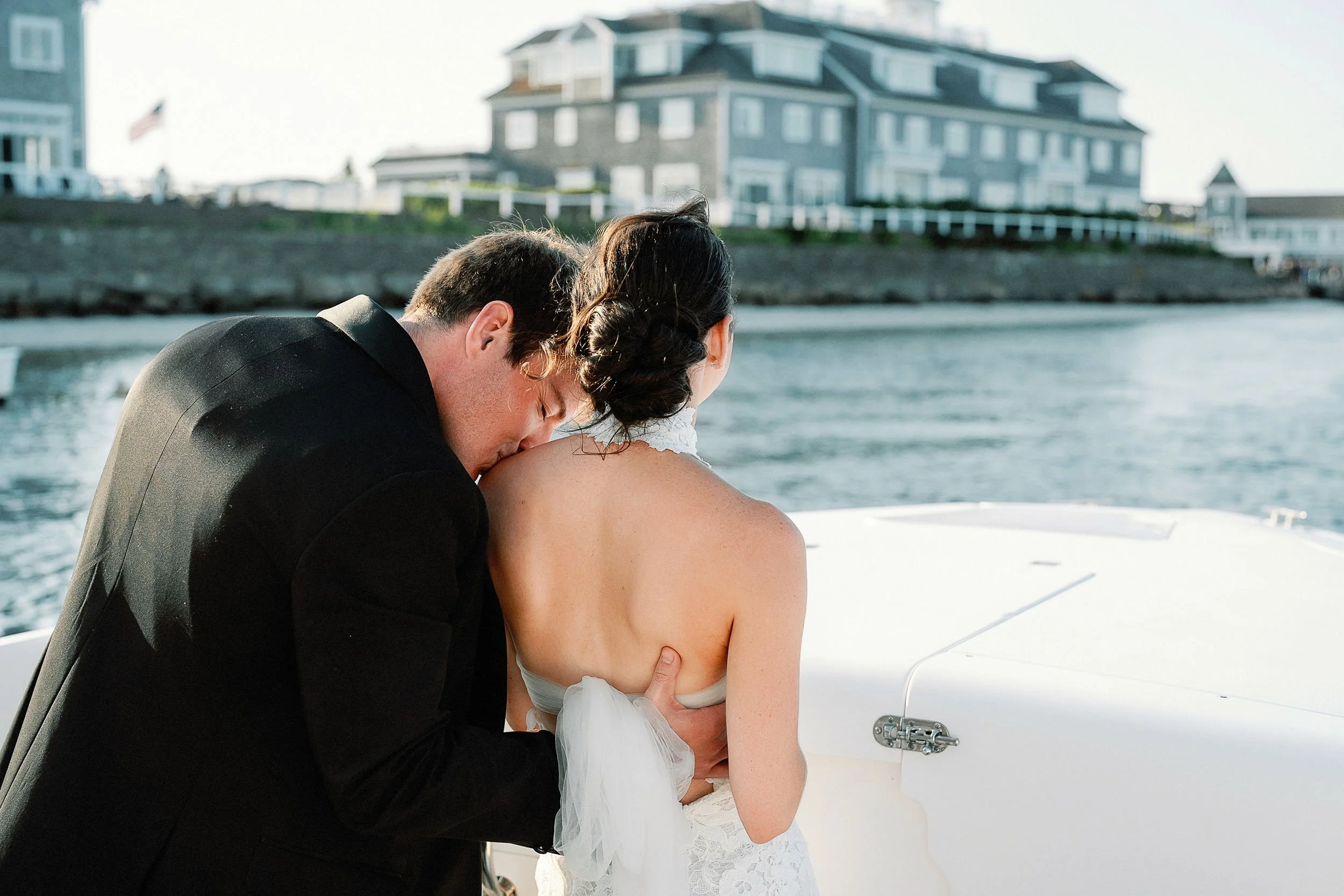 A groom kissing a bride on the neck on a boat with houses along a shoreline in the background.