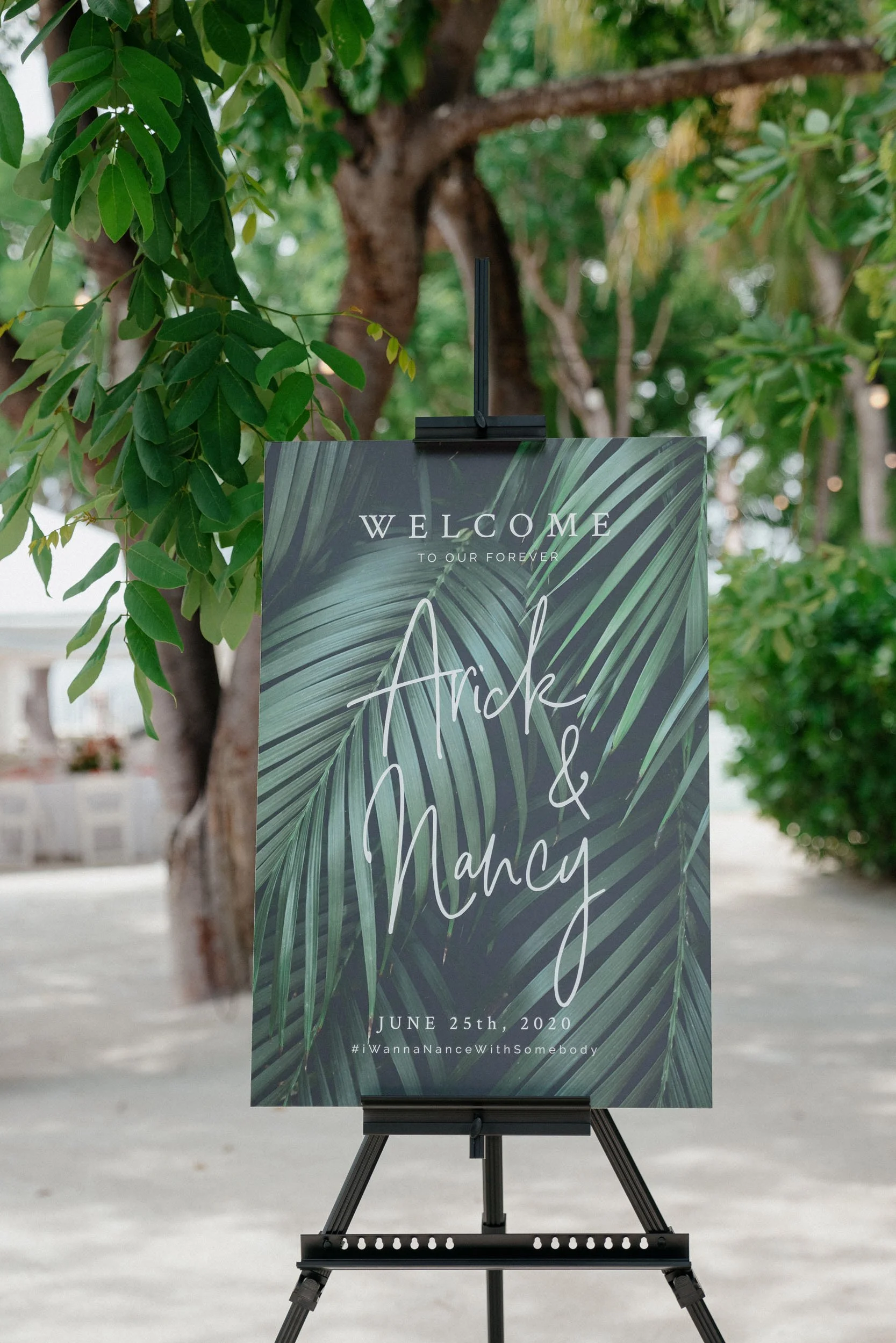 Wedding welcome sign with tropical palm leaves background, indicating a wedding event for friends Aride and Nancy on June 25th, 2020, displayed on an easel outdoors.