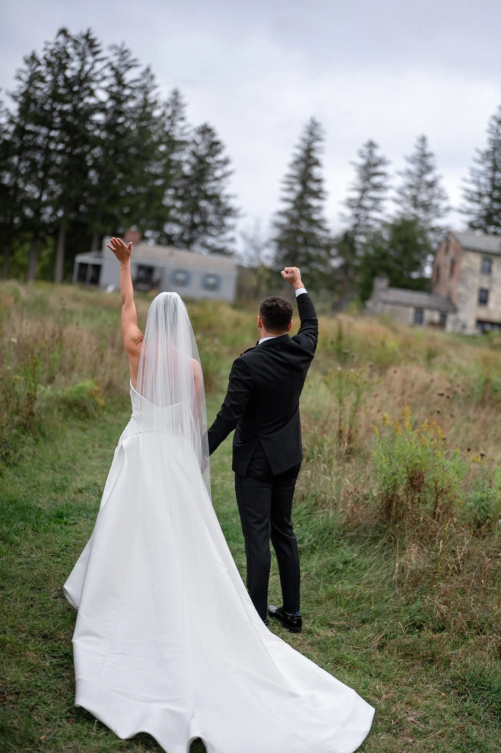 Bride and groom walking on a grassy path outdoors, with the bride raising her left arm in celebration and both wearing formal wedding attire.