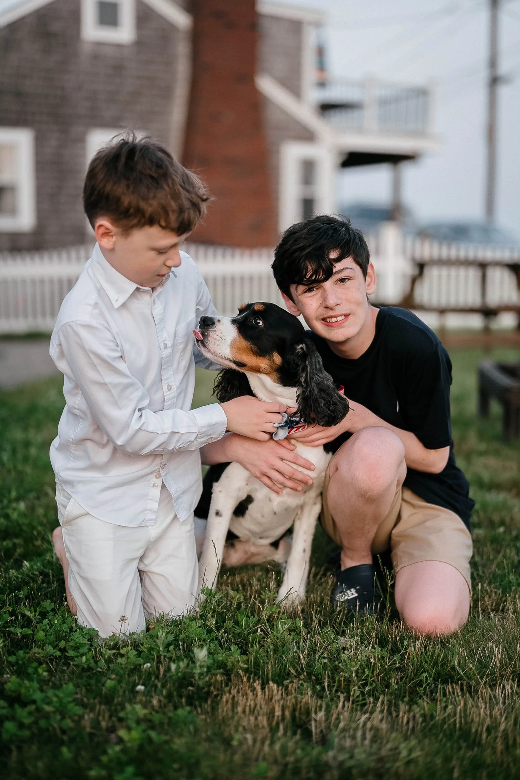 Two boys and a dog in a backyard, one boy wearing a white shirt and the other in a black shirt, with the dog in the center.