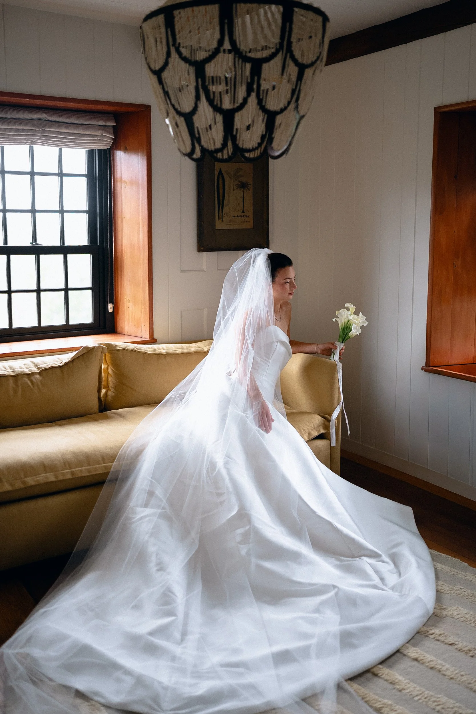 Brunette bride in white wedding gown and veil sitting on a yellow sofa holding a bouquet of calla lilies inside a cozy room with wooden window frames and a decorative lamp overhead.