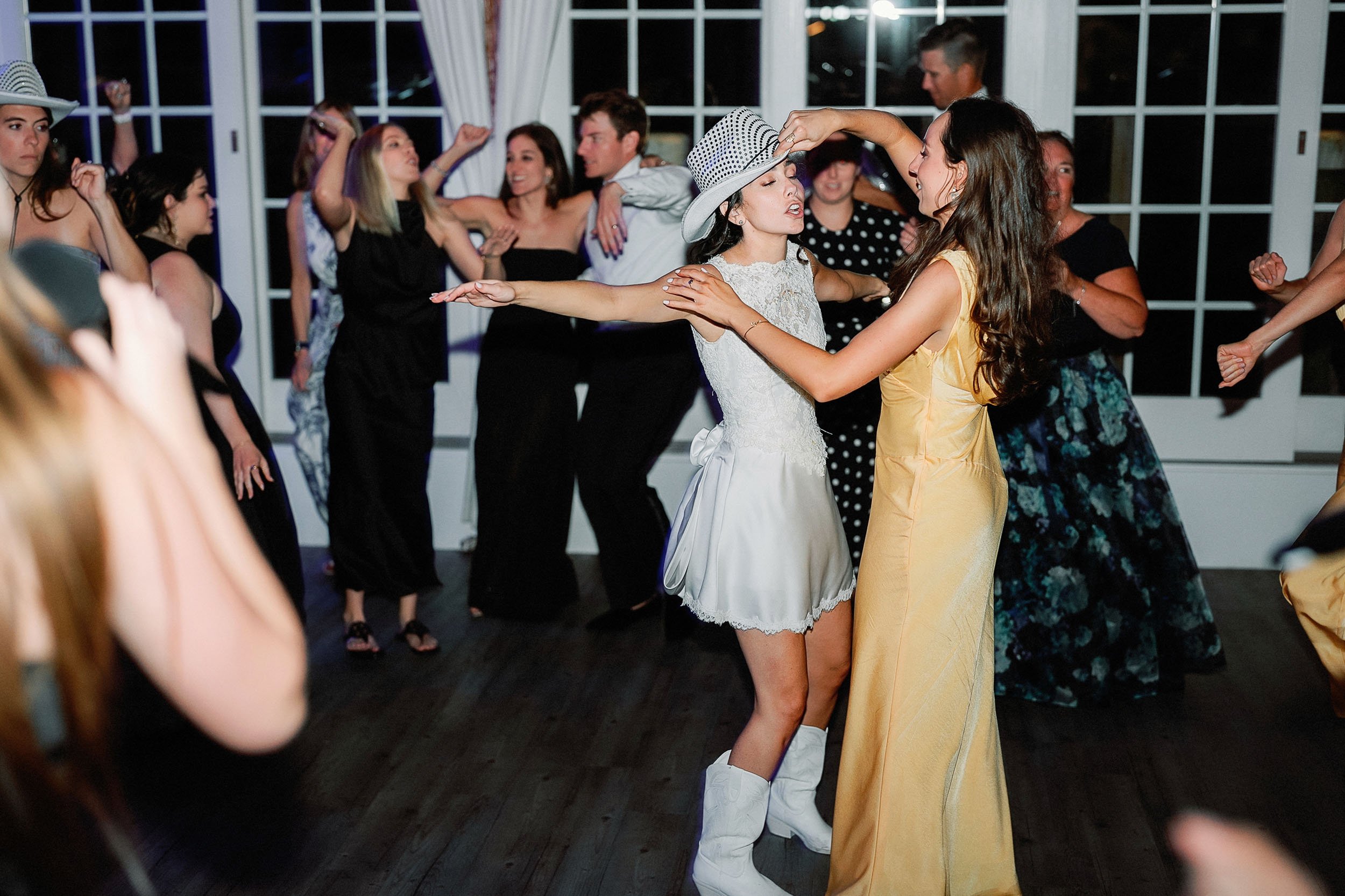 People dancing at a party or celebration with a woman in a white dress and cowboy hat and another woman in a yellow dress dancing together. Others dancing in the background.