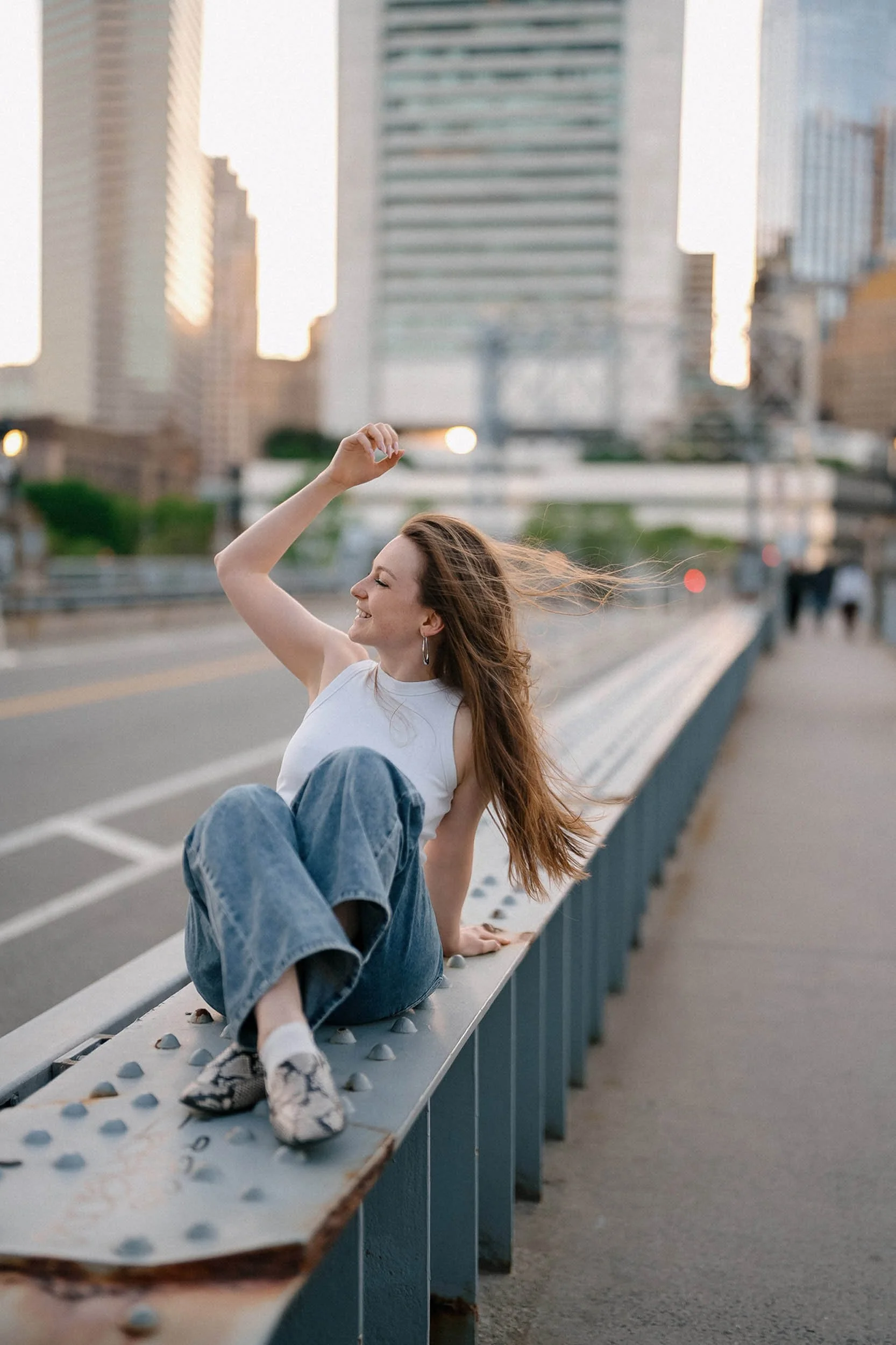 A woman with long, wavy hair is sitting on a metal barrier on a city street. She is wearing a white sleeveless top and baggy jeans, smiling and looking to her left as her hair flows in the wind during sunset.