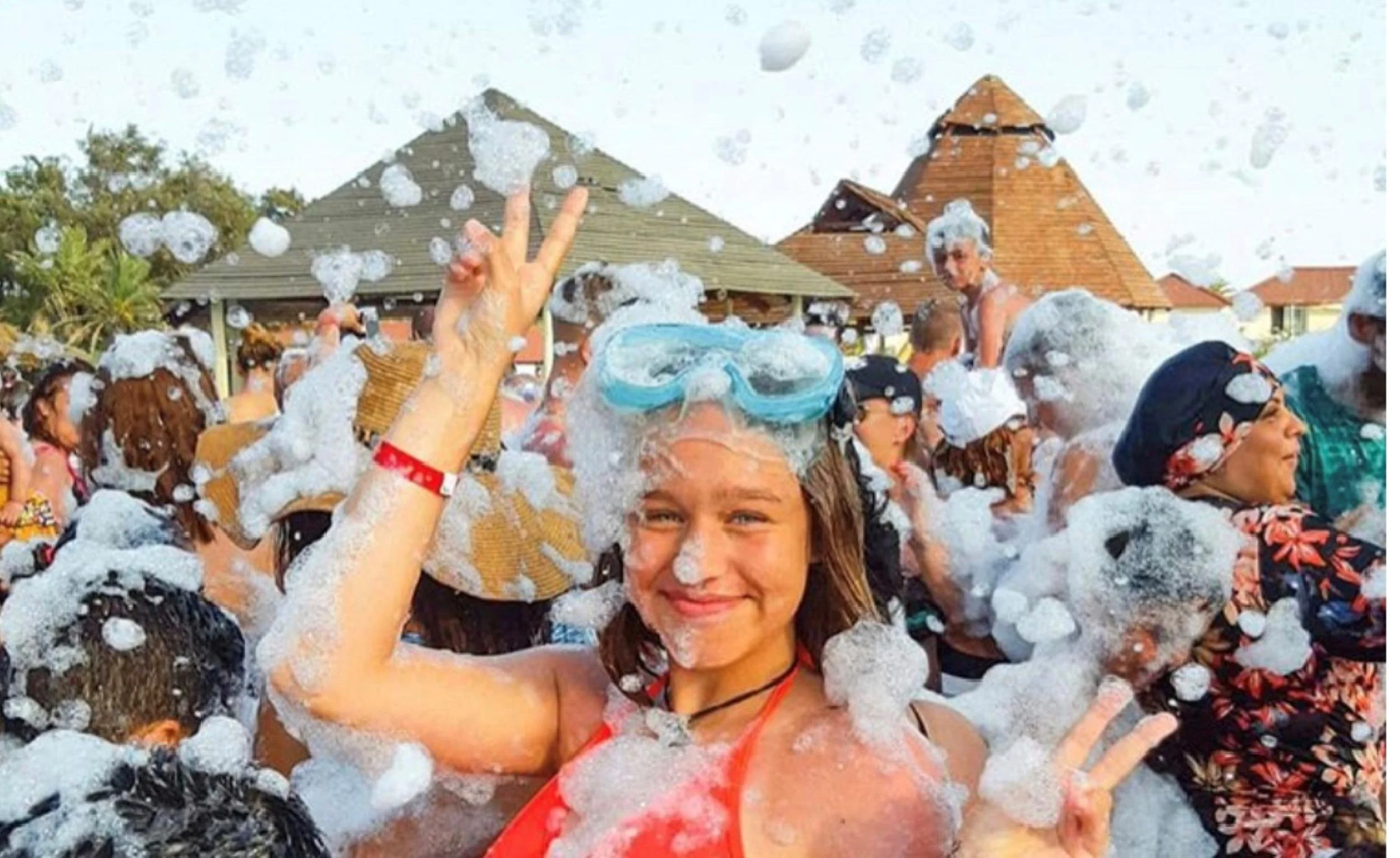 A smiling teen girl wearing blue goggles flashes a peace sign covered in foam at a lively outdoor foam party event in San Jose, California.