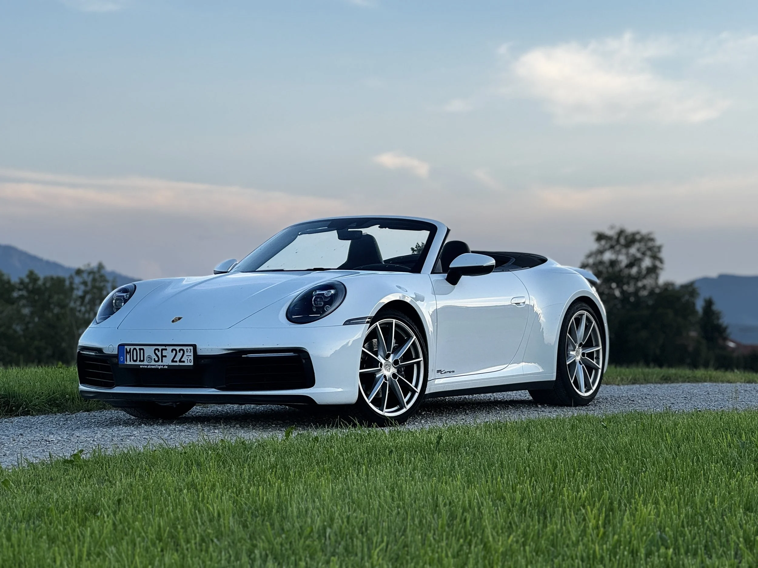 Ein weißer Porsche 911 Cabrio auf einem grasbewachsenen Feld bei Sonnenuntergang.
